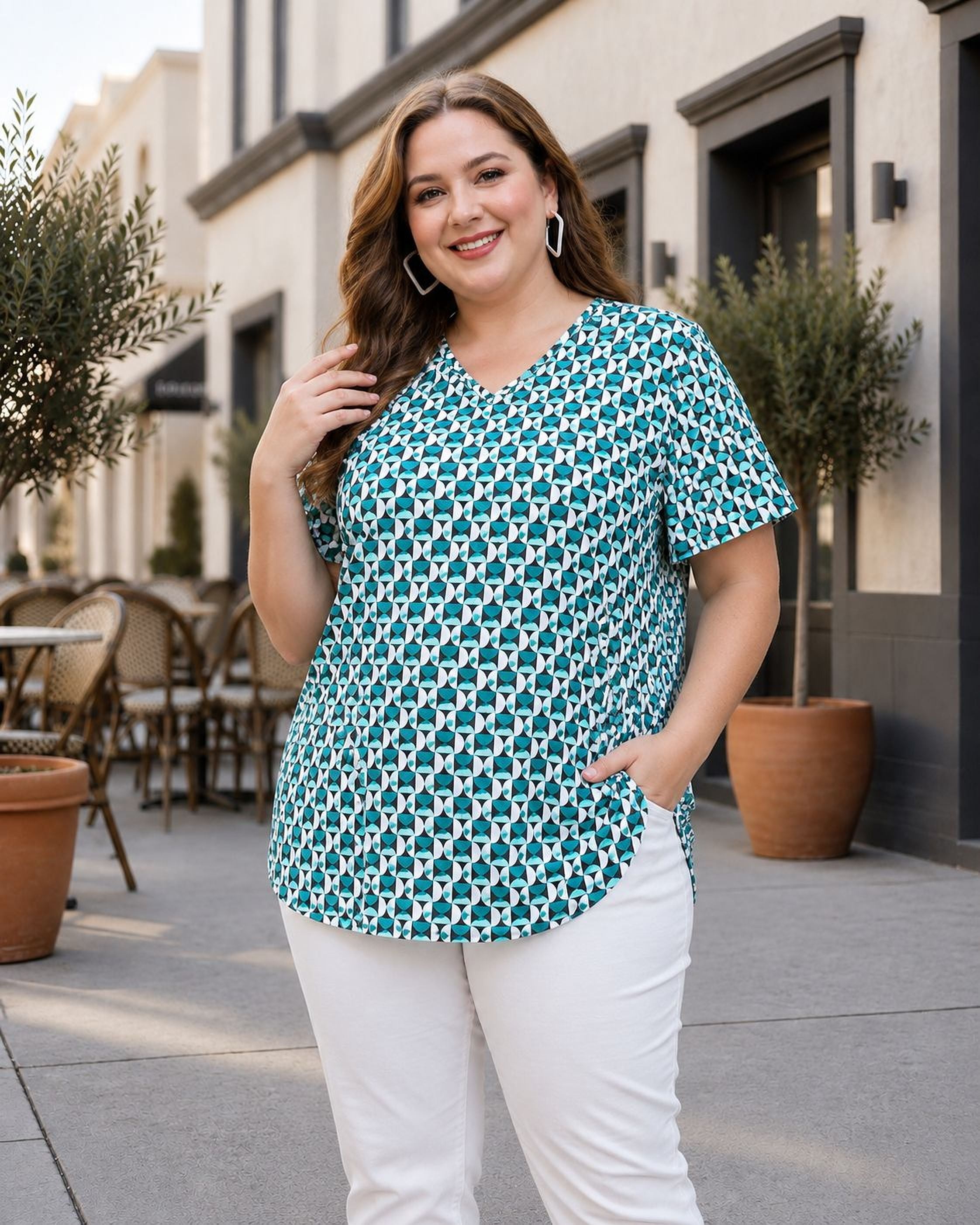 A woman wearing a teal and white patterned blouse and white pants is standing on a sidewalk in front of a building with potted plants.