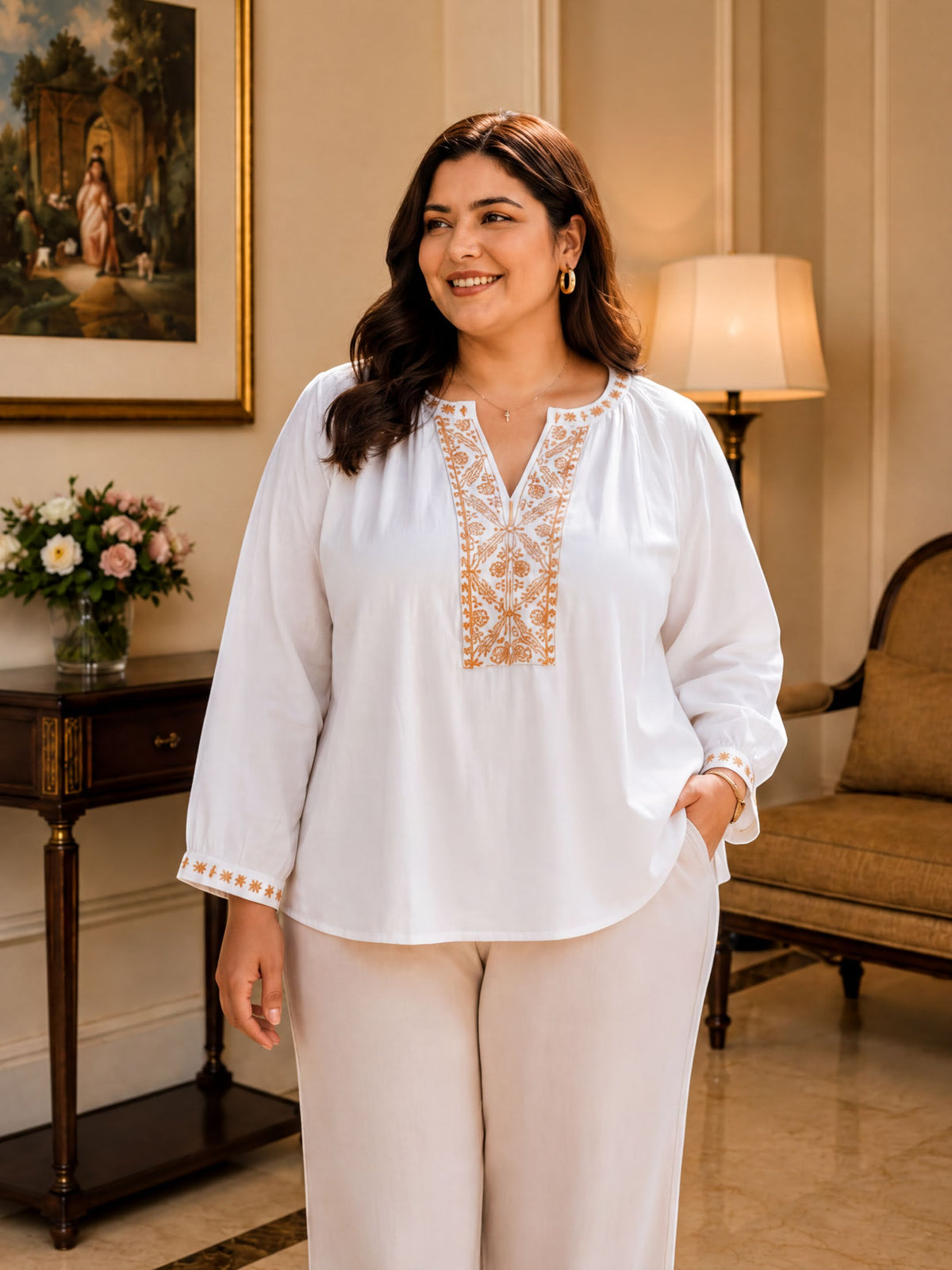 A woman in a white blouse with gold embroidery stands in a room with a painting on the wall, a lamp, and a vase of flowers.