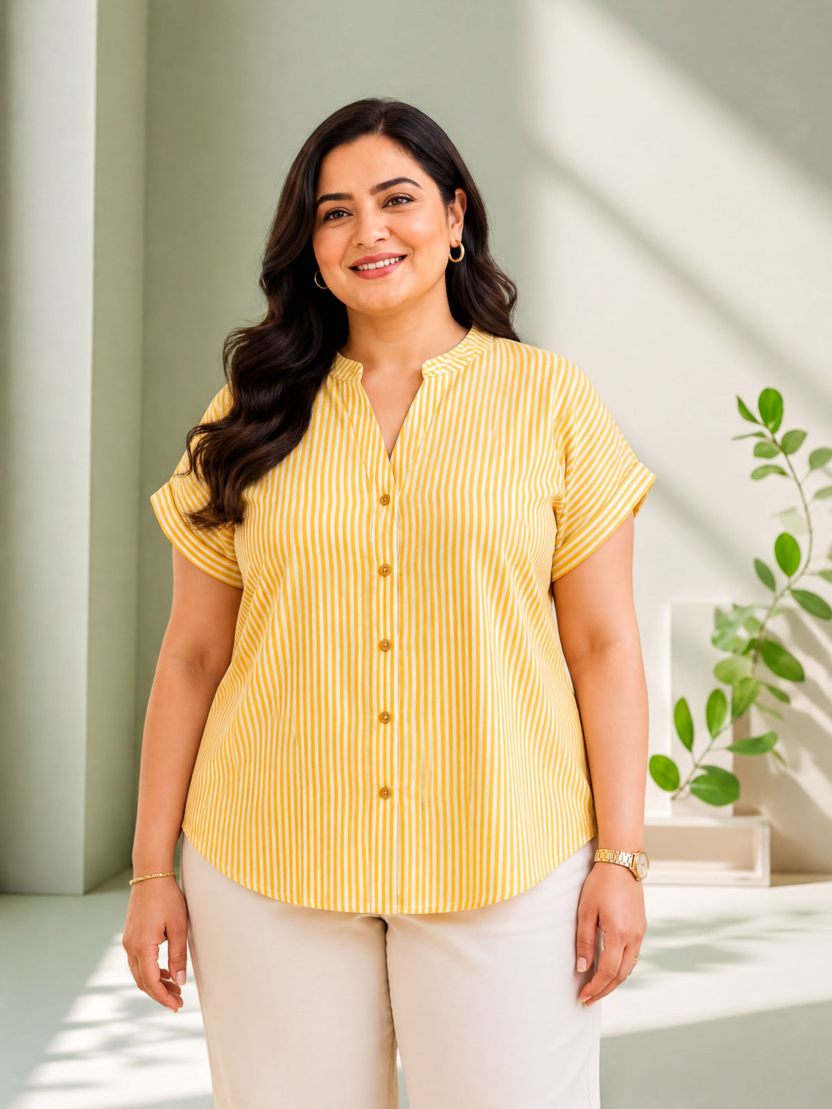 A woman with long dark hair is smiling and wearing a yellow and white striped blouse and white pants.