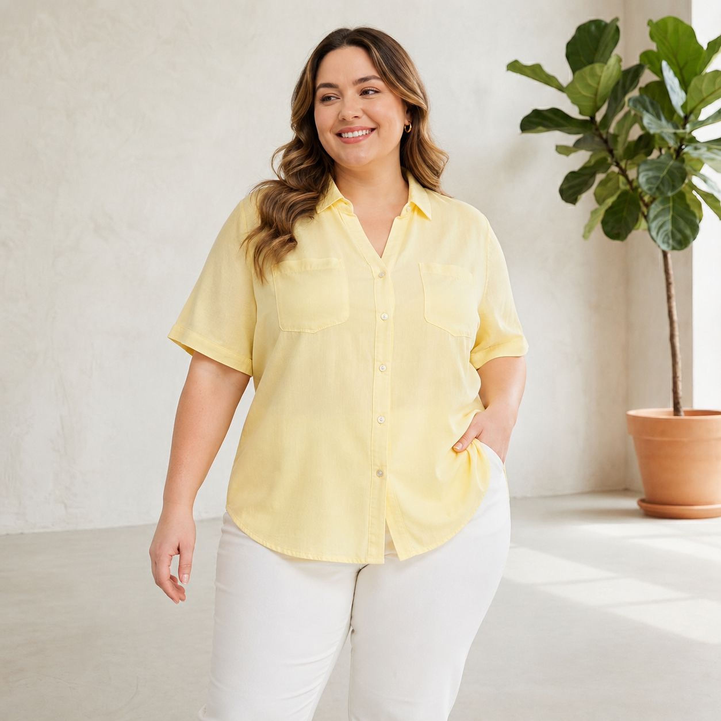 A woman wearing a yellow shirt and white pants is smiling and posing in a room with a potted plant.