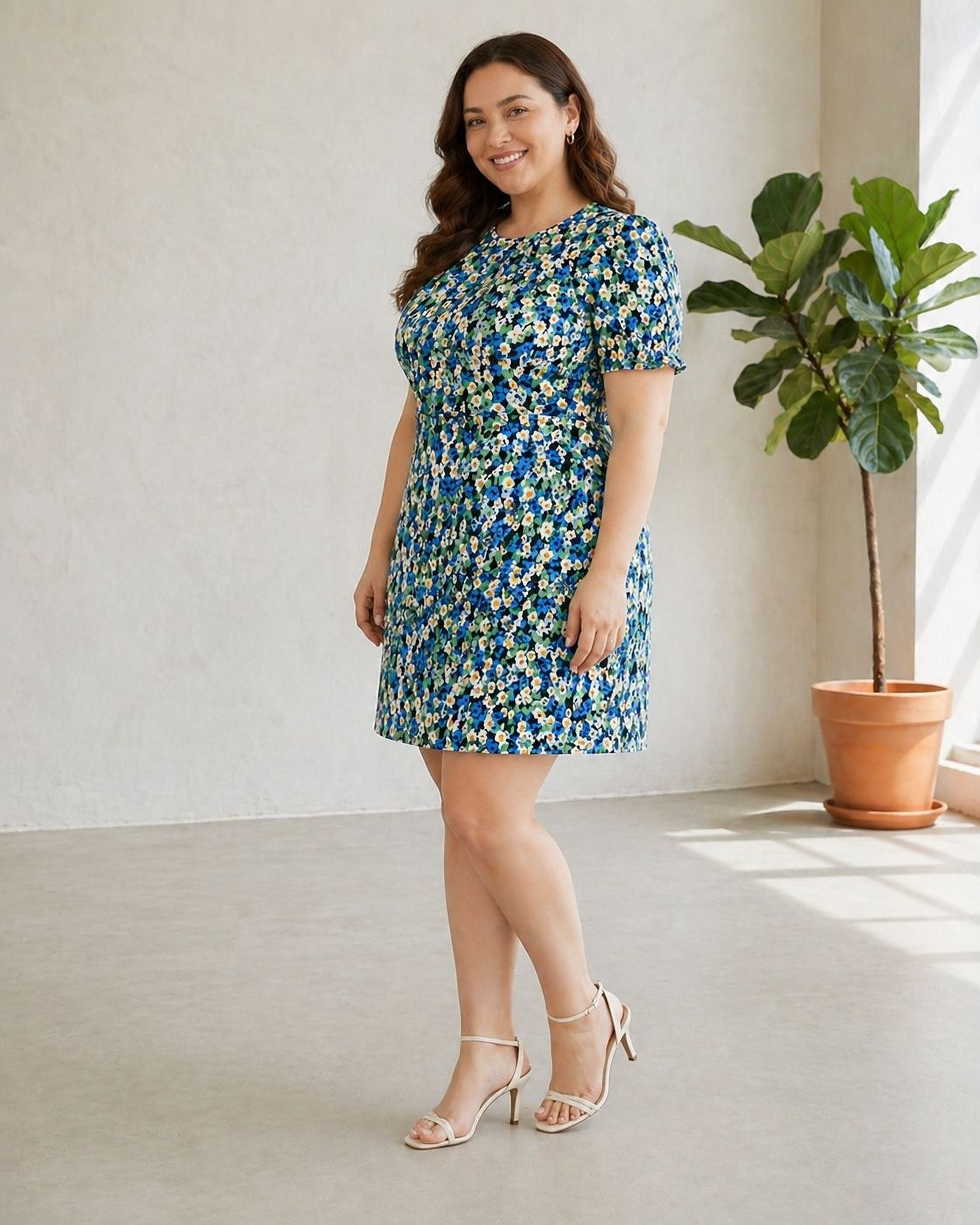 A woman wearing a blue and green floral print dress and high heels stands in a room with a potted plant.