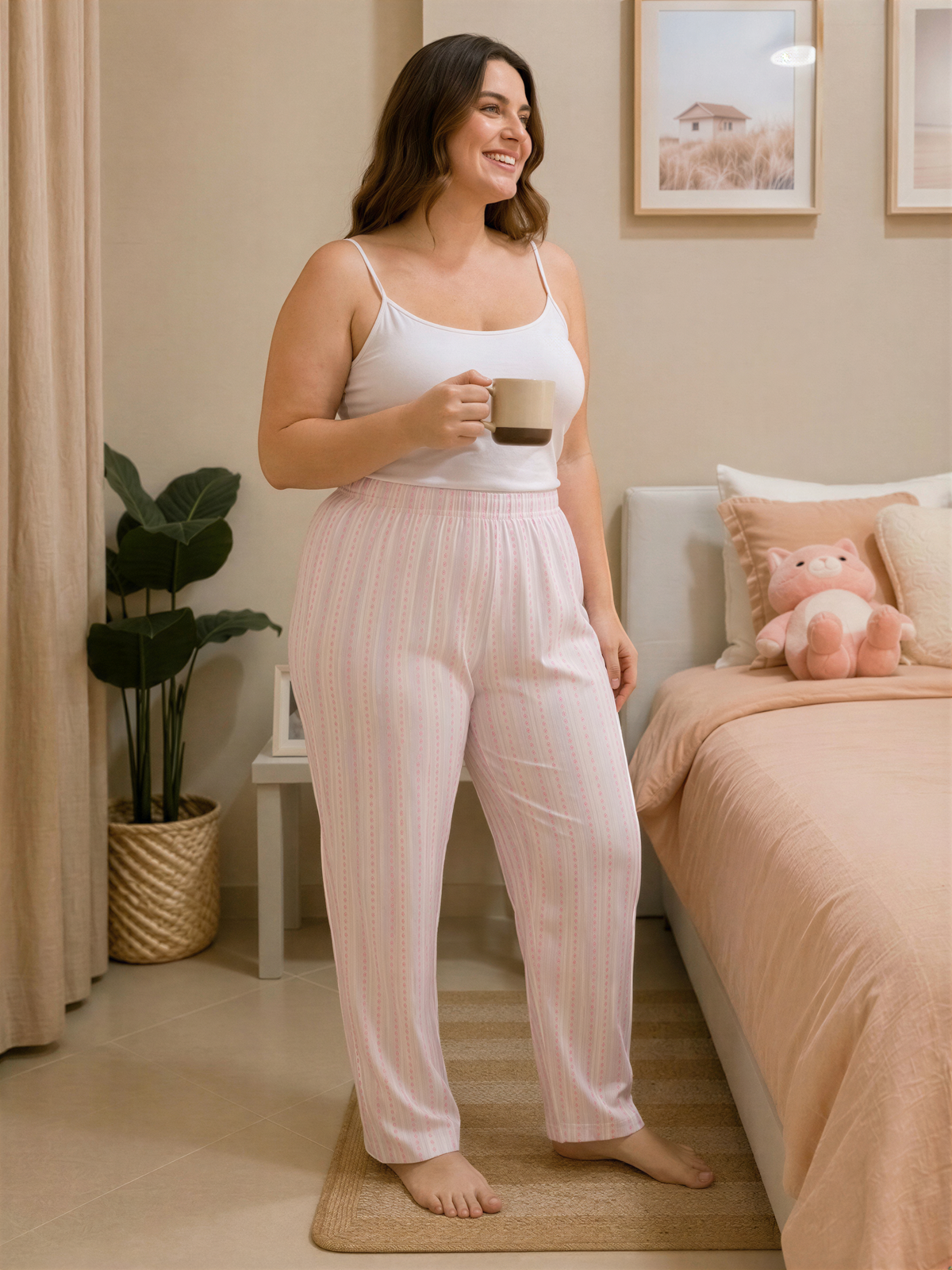 A woman in a white tank top and striped pajama pants stands in a bedroom, holding a coffee mug and smiling.
