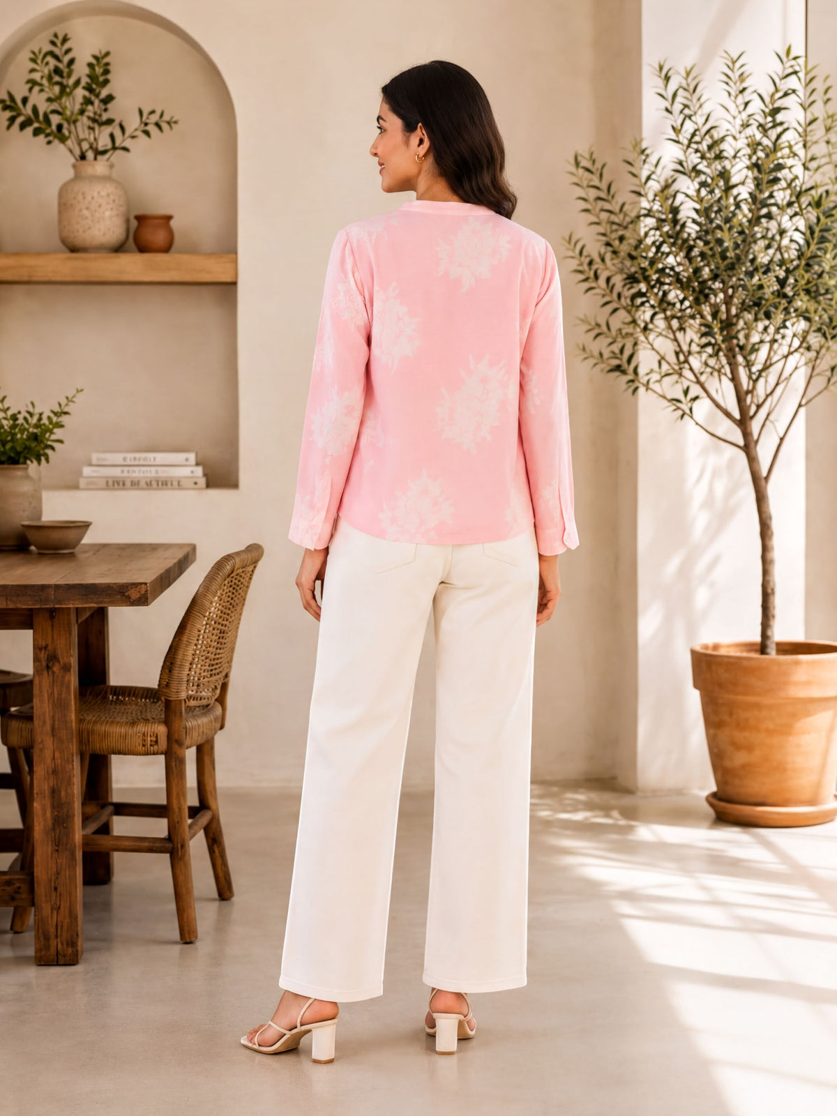 A woman wearing a pink blouse and white pants stands in a room with a wooden table and chairs, a potted plant, and a shelf with decorative items.