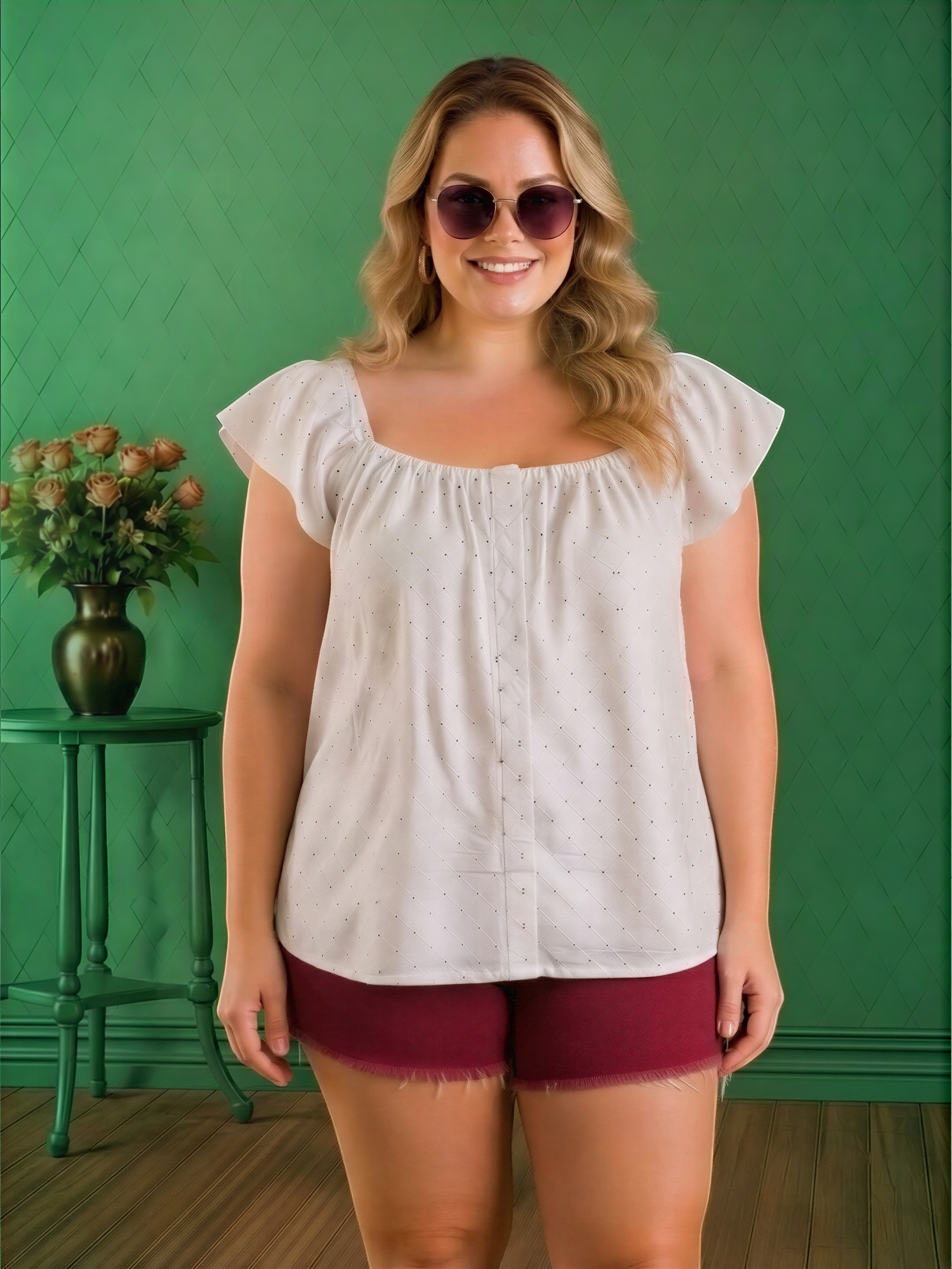 A woman with blonde hair and sunglasses is standing in front of a green wall, wearing a white blouse with a ruffled collar and maroon shorts.