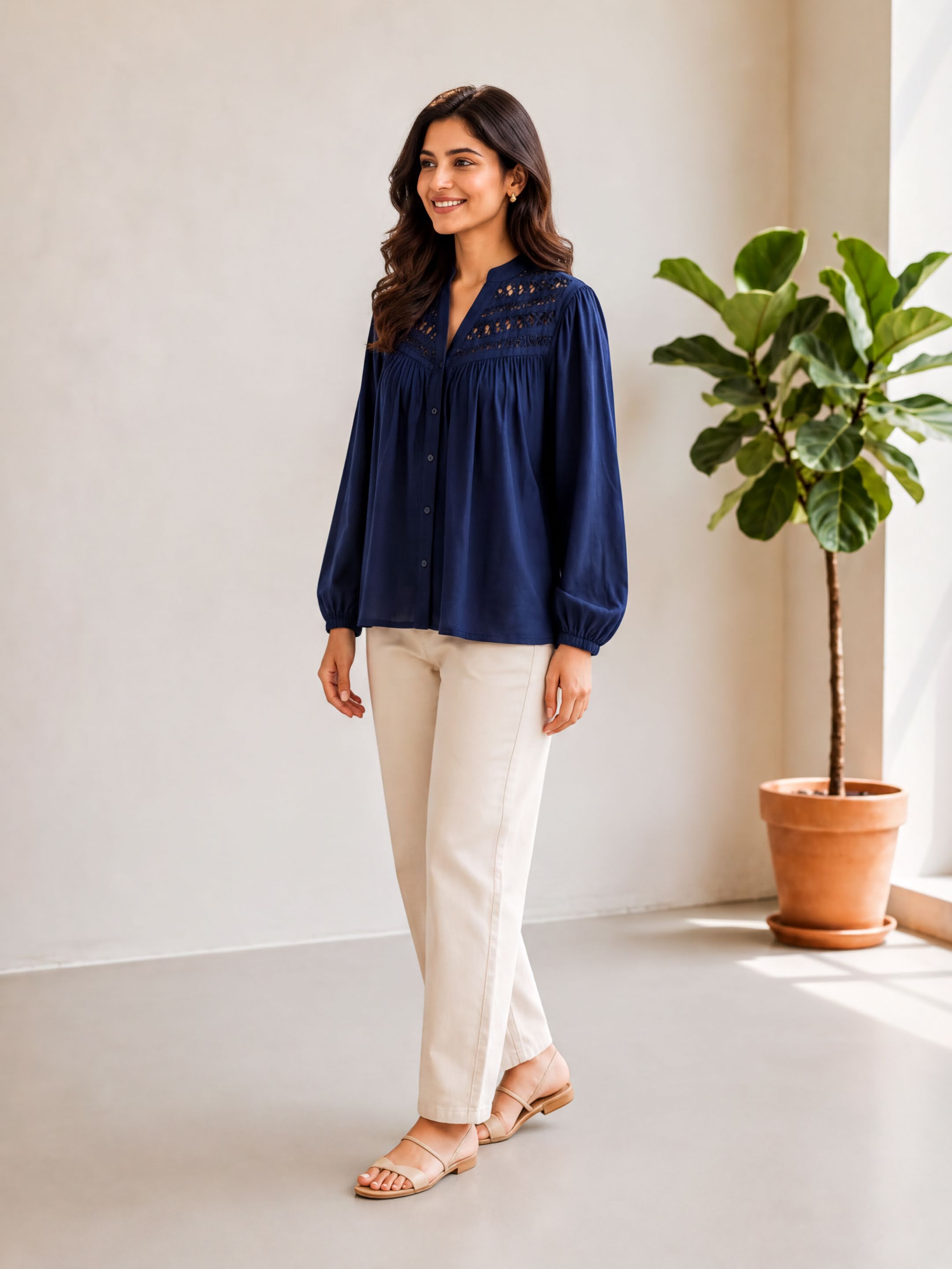 A woman in a navy blue blouse and beige pants stands in a room with a potted plant to her right.