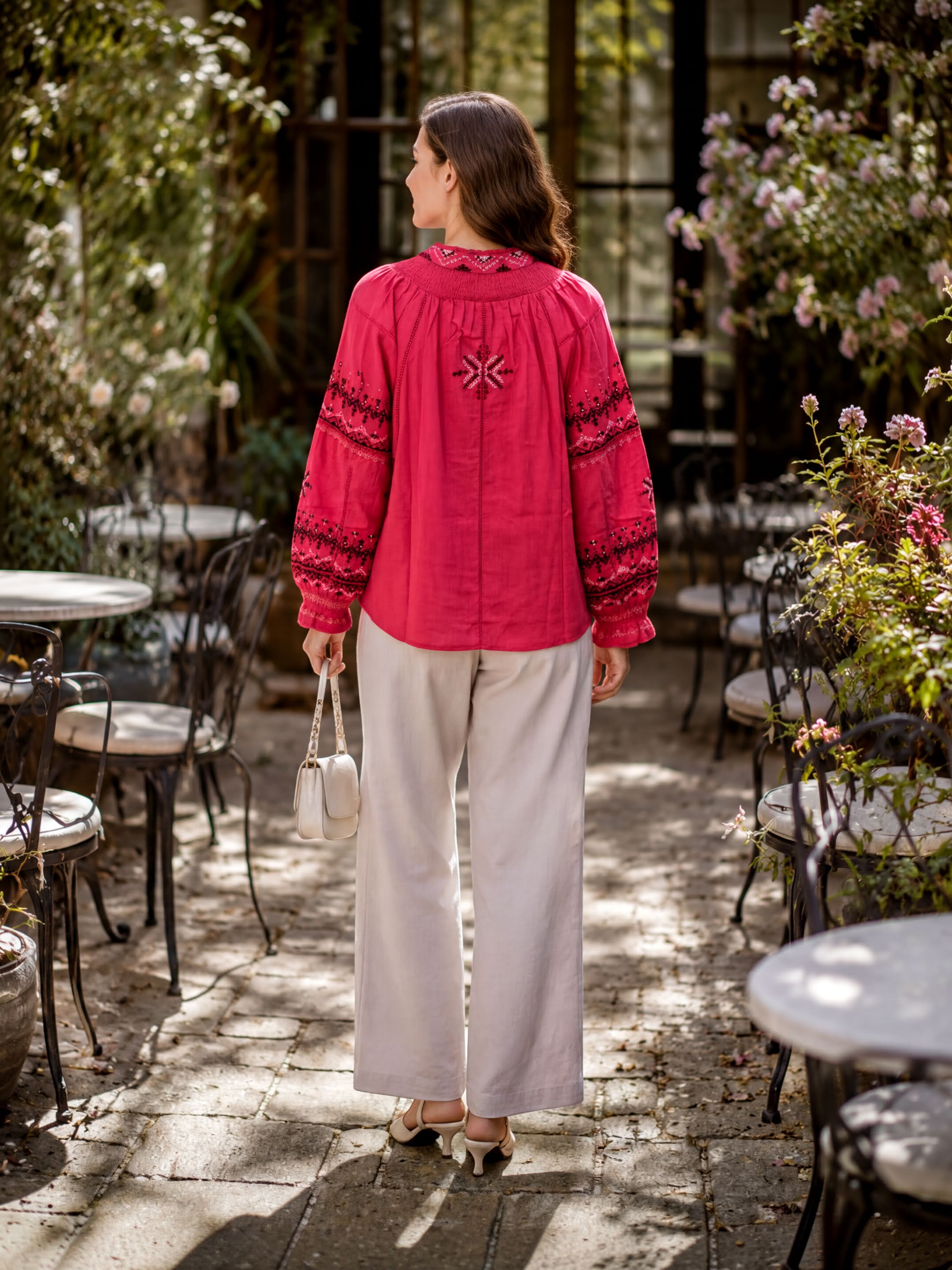 A woman in a red blouse and white pants stands in a garden, facing away from the camera.