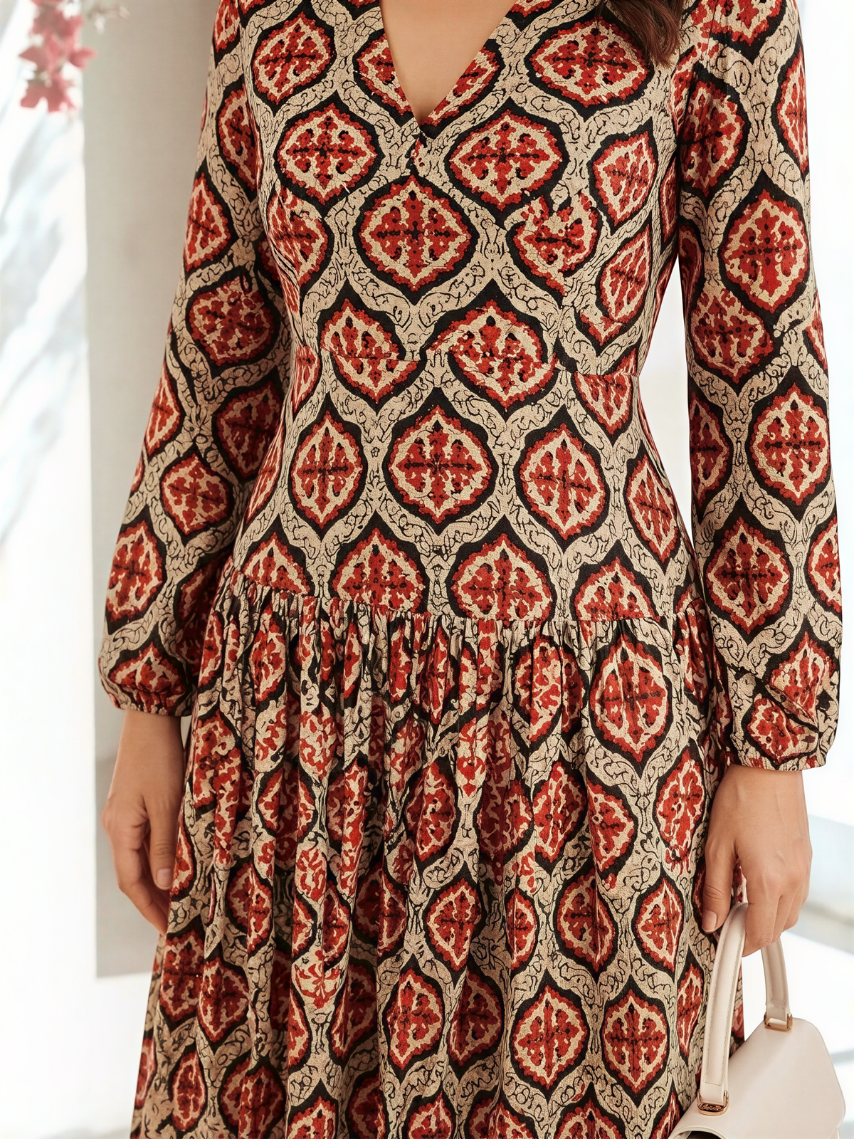 A woman wearing a long, patterned dress with a v-neck and a flared skirt. She is holding a white purse in her hand.