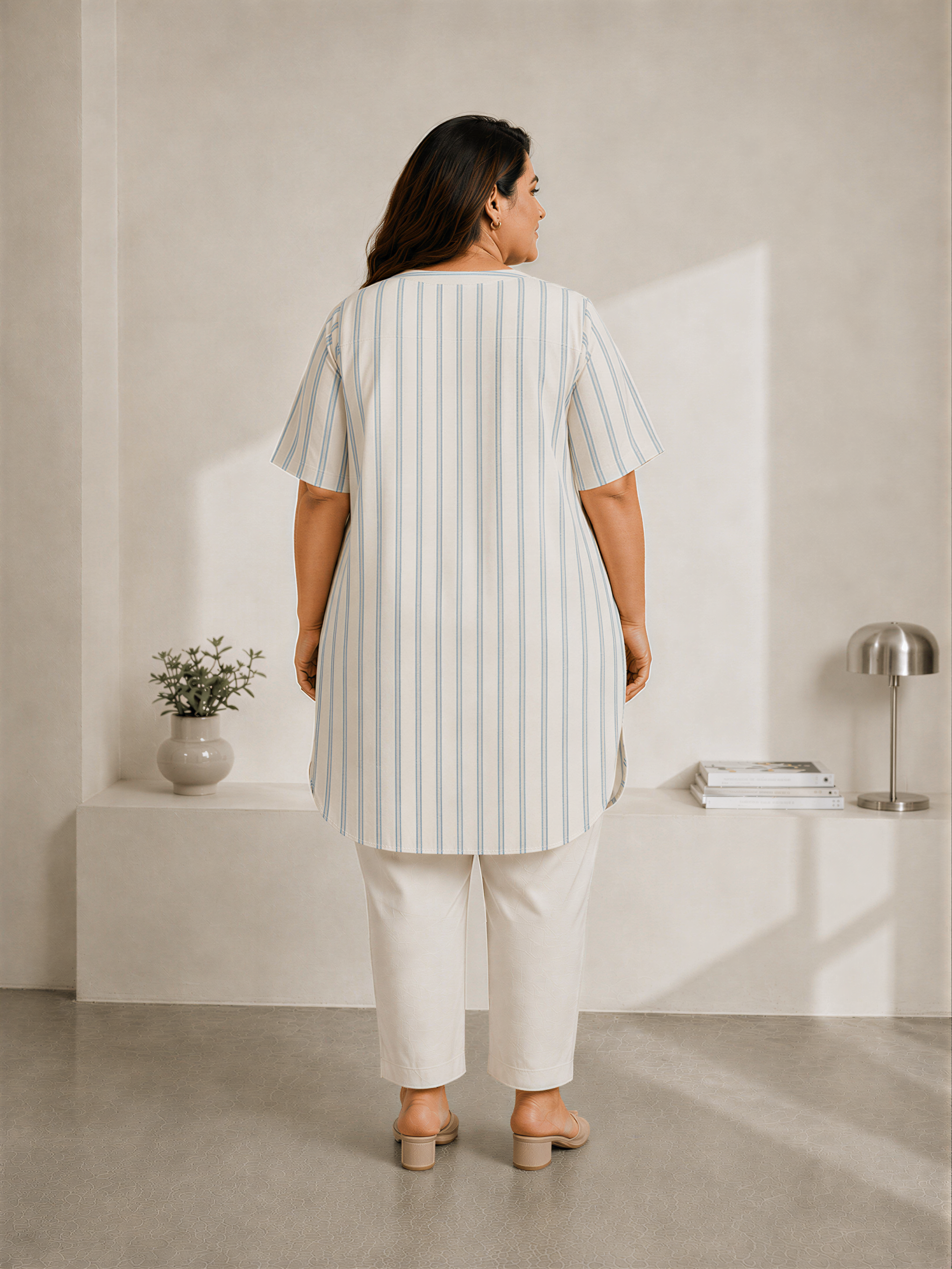 A woman wearing a striped shirt and white pants stands in a minimalist room with a white wall, a small plant, and a lamp.