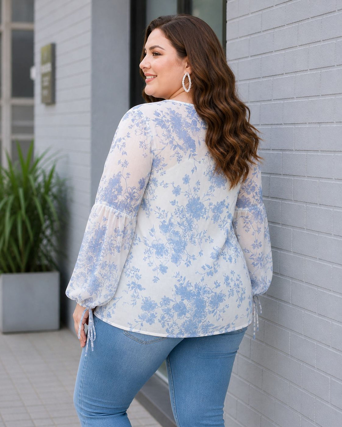 A woman with long brown hair is standing in front of a gray building, smiling and looking to her left. She is wearing a white blouse with blue floral patterns and blue jeans.
