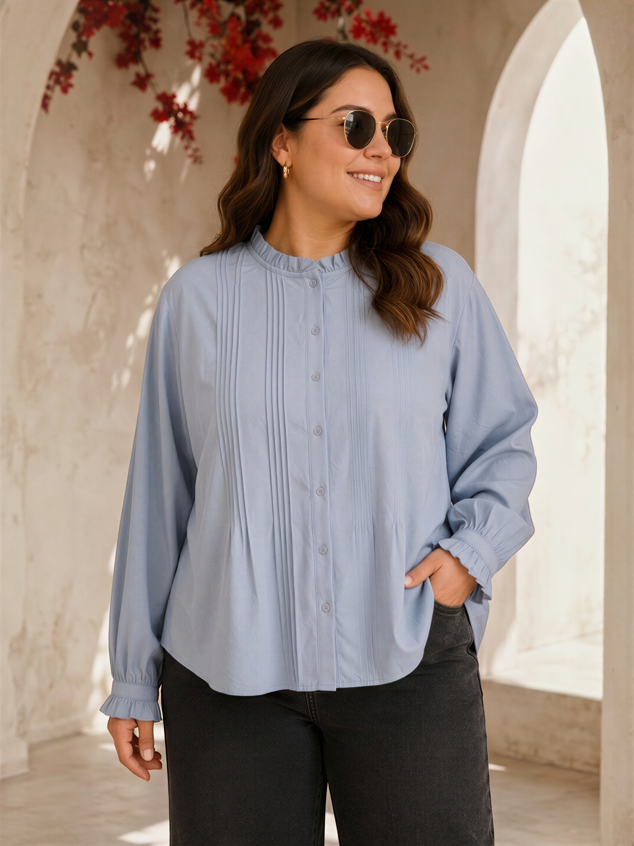 A woman wearing a light blue button-up shirt and sunglasses stands in front of a white archway with red flowers hanging from it.