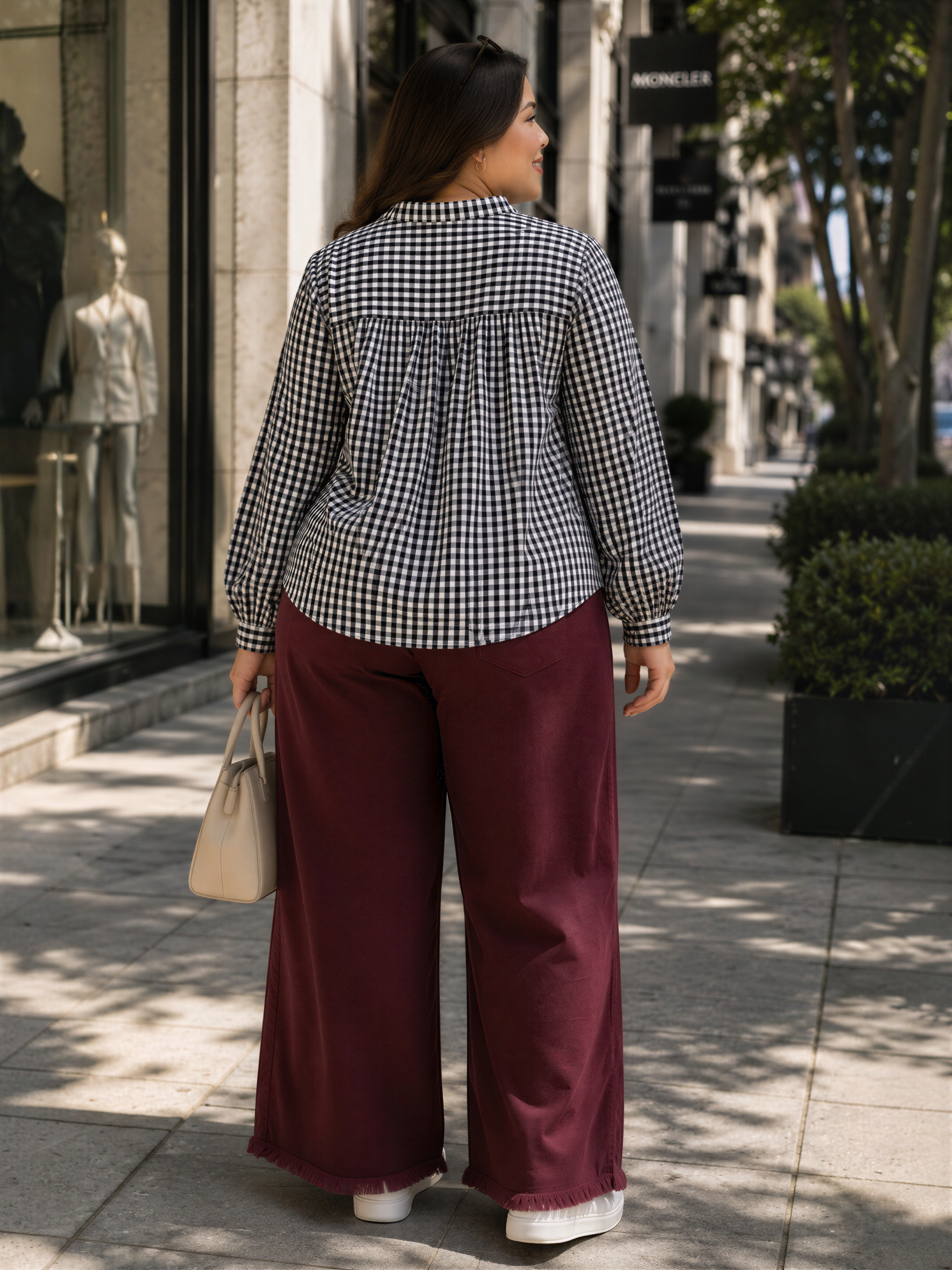A woman wearing a black and white checkered blouse and burgundy wide-leg pants is standing on a sidewalk, looking away from the camera.