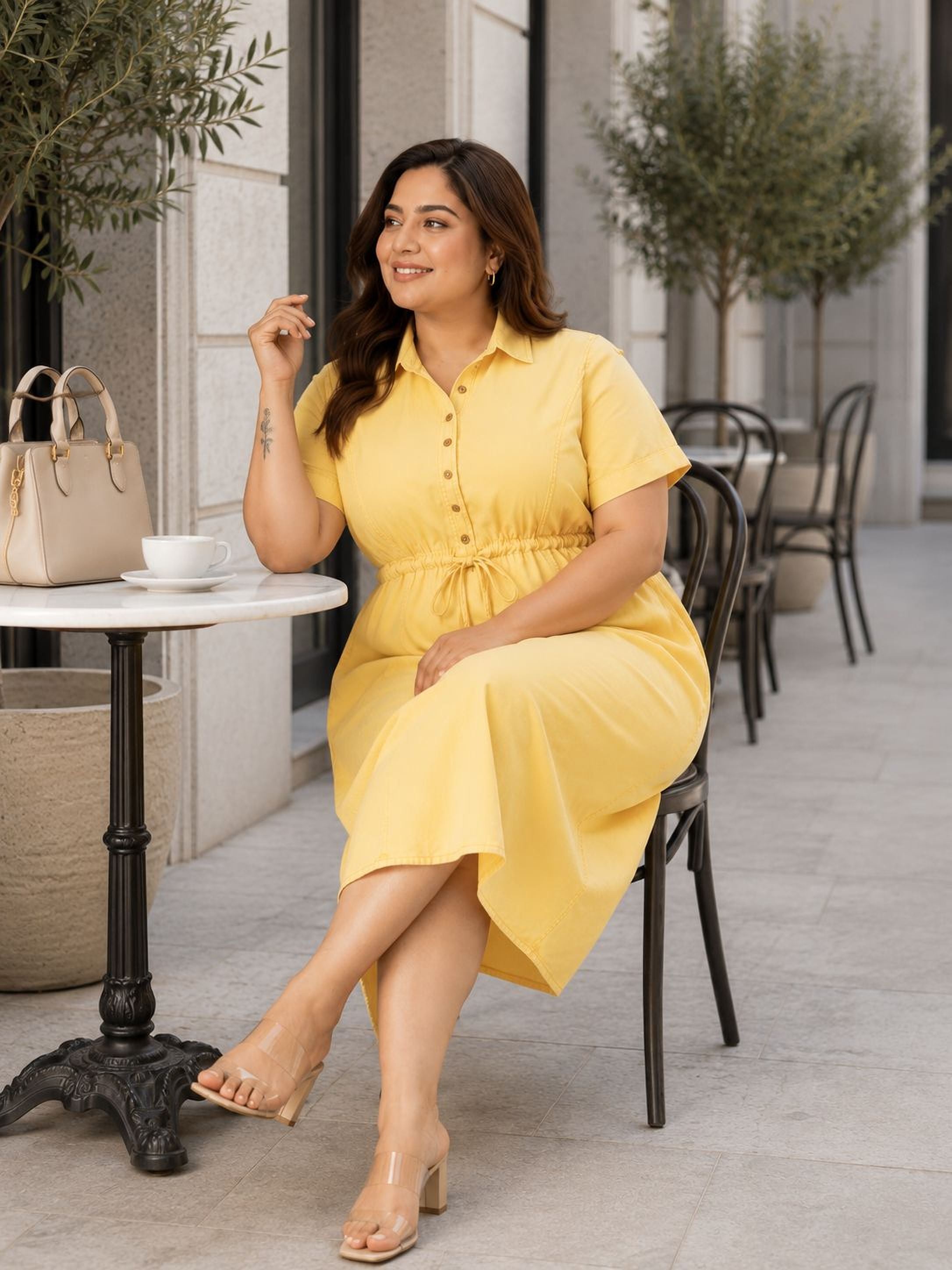 A woman in a yellow dress is sitting at a table outside, smiling and holding a cup of coffee.