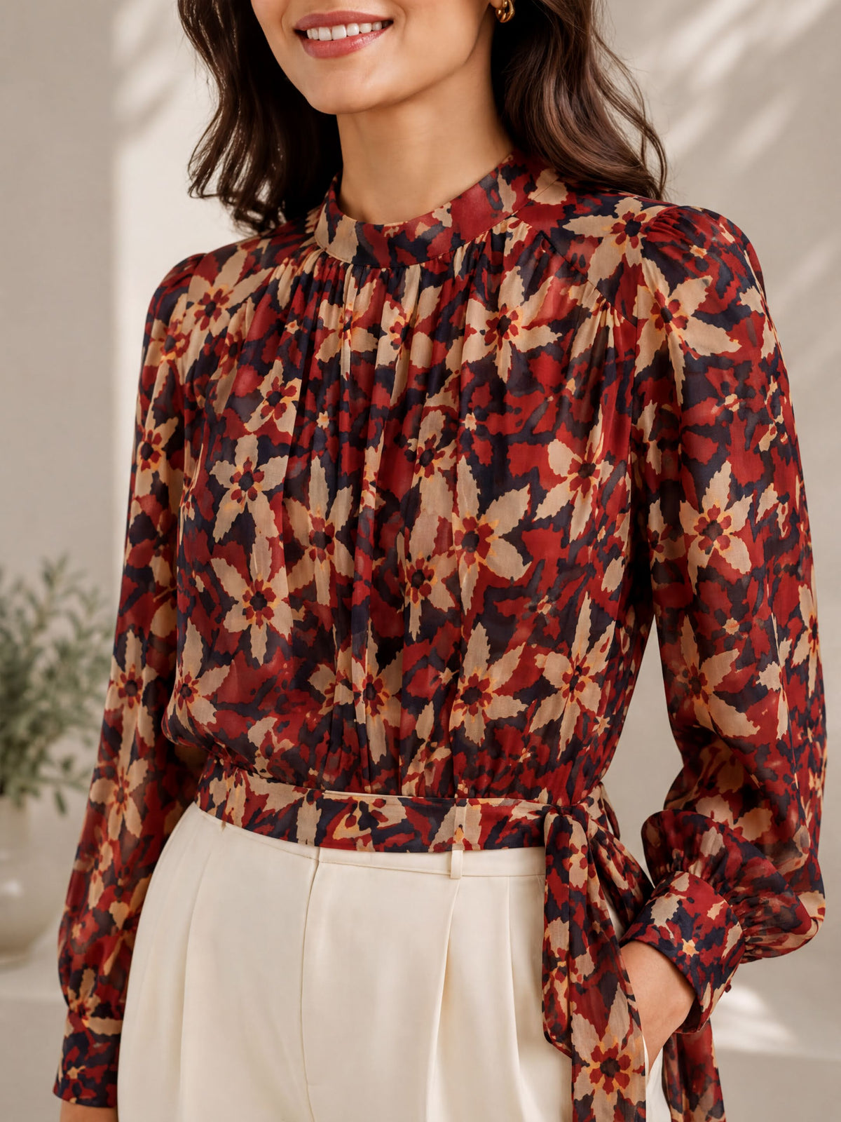A woman wearing a red and black floral blouse with a high collar and long sleeves, paired with white pants.