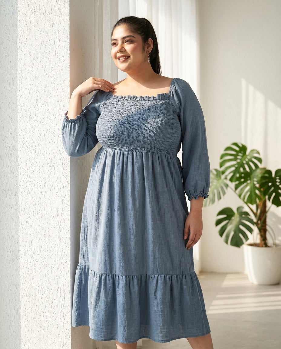 Smiling woman in blue smocked midi dress with long sleeves, standing by a window indoors