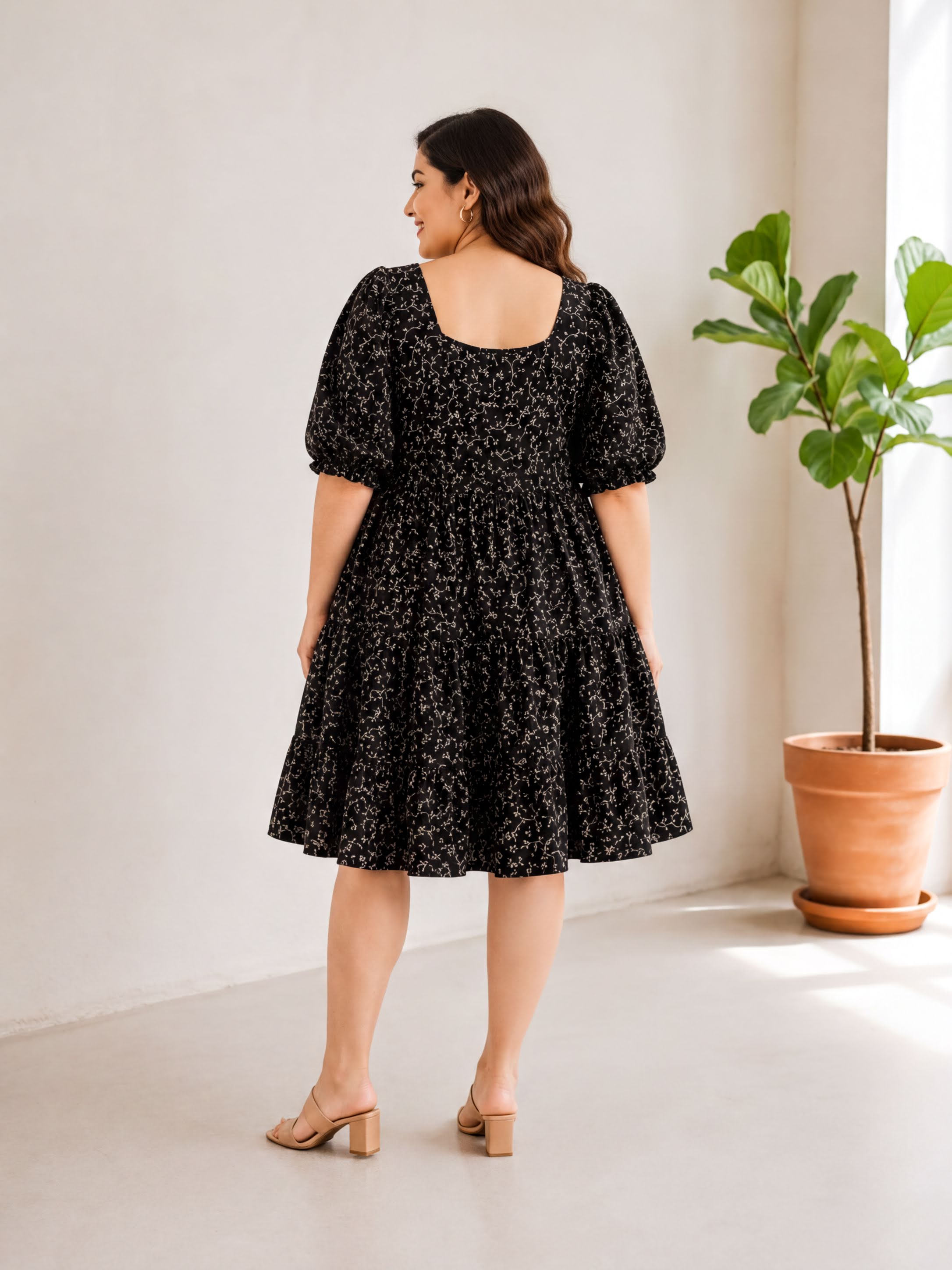 A woman wearing a black dress with a floral pattern stands in a room with a potted plant.