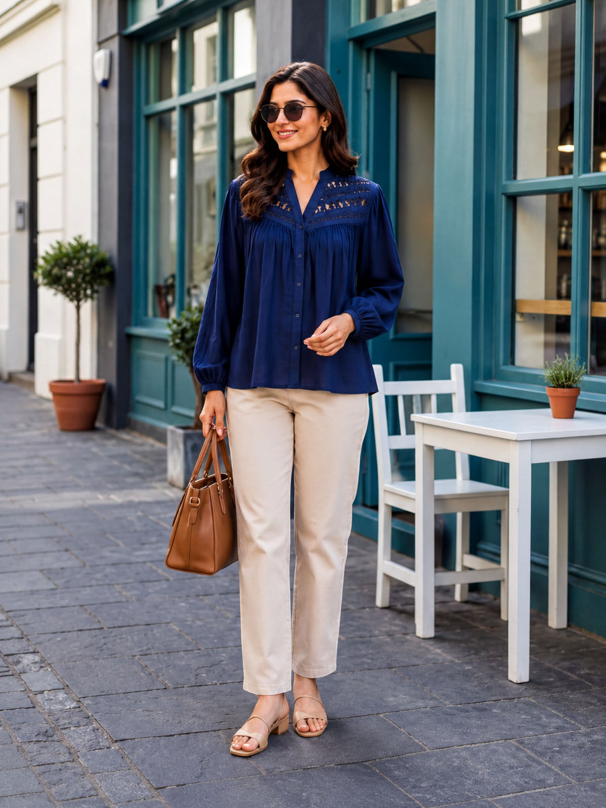 A woman wearing a navy blue blouse and beige pants is walking on a sidewalk, carrying a brown purse and smiling.