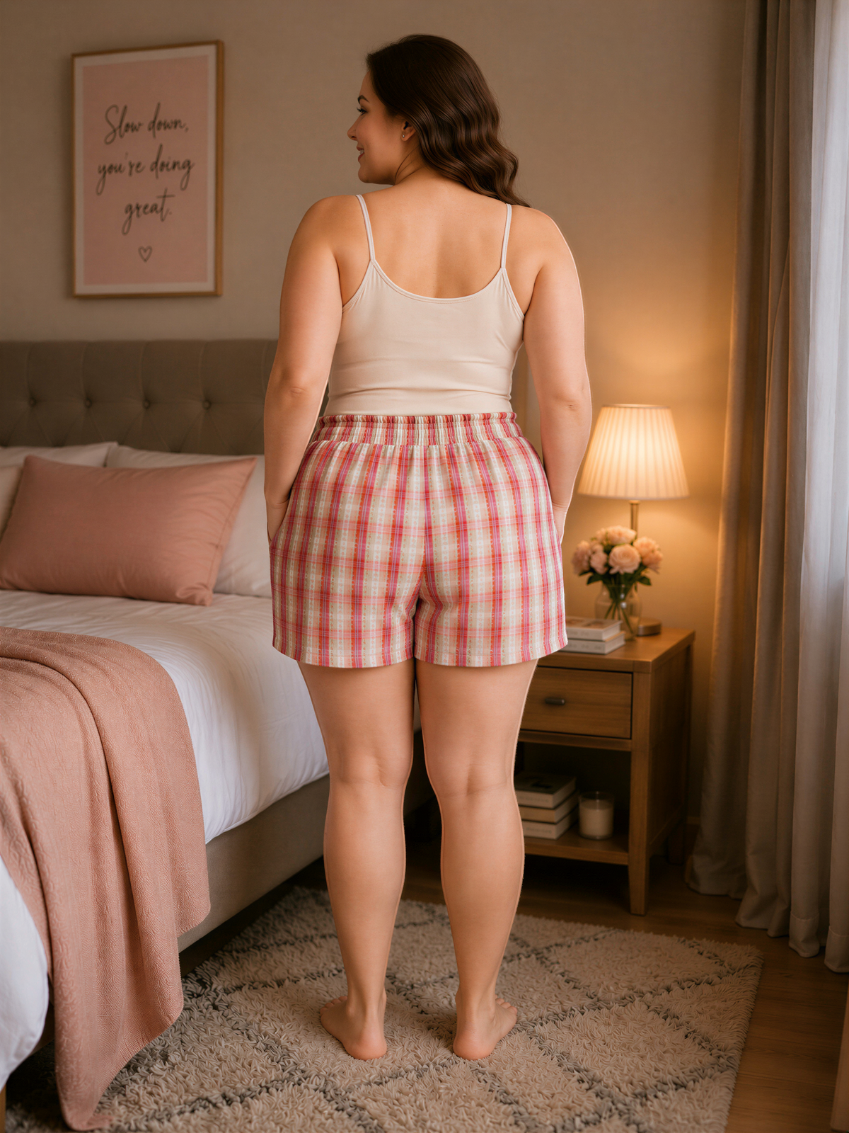 A woman stands in a bedroom, wearing a white tank top and red and white plaid shorts. She is positioned in front of a bed with a pink blanket and a nightstand with a lamp. The wall behind her has a framed quote that reads "Slow down, you're doing great."