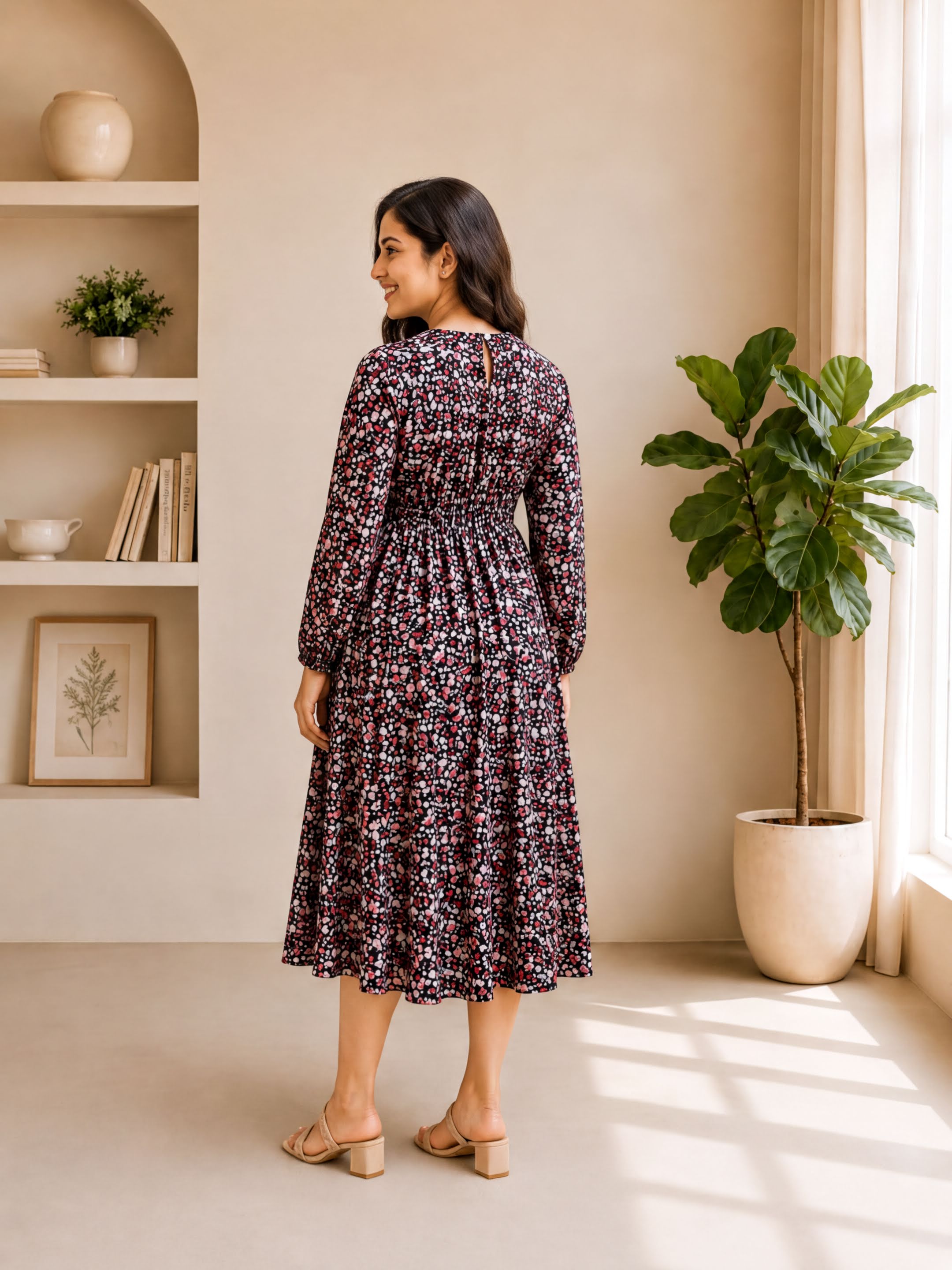 A woman wearing a long, floral print dress stands in a room with a window, plants, and shelves.