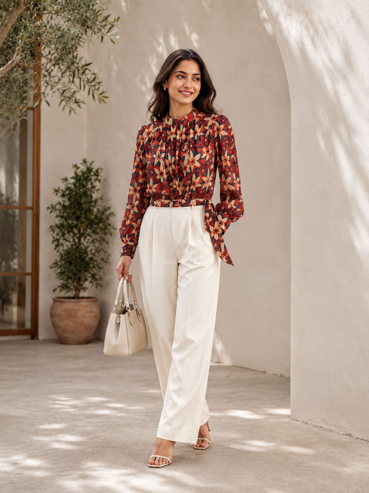 A woman wearing a red and black floral blouse and white pants, standing in front of a white wall with a potted plant to her left. She is holding a beige handbag and smiling at the camera.