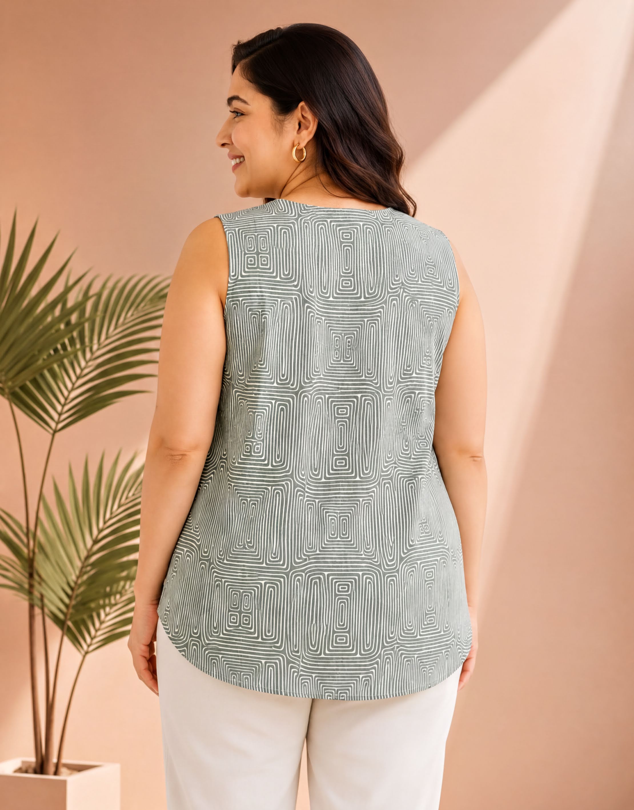 A woman wearing a sleeveless top with a patterned design, standing in front of a palm plant and a pink wall.