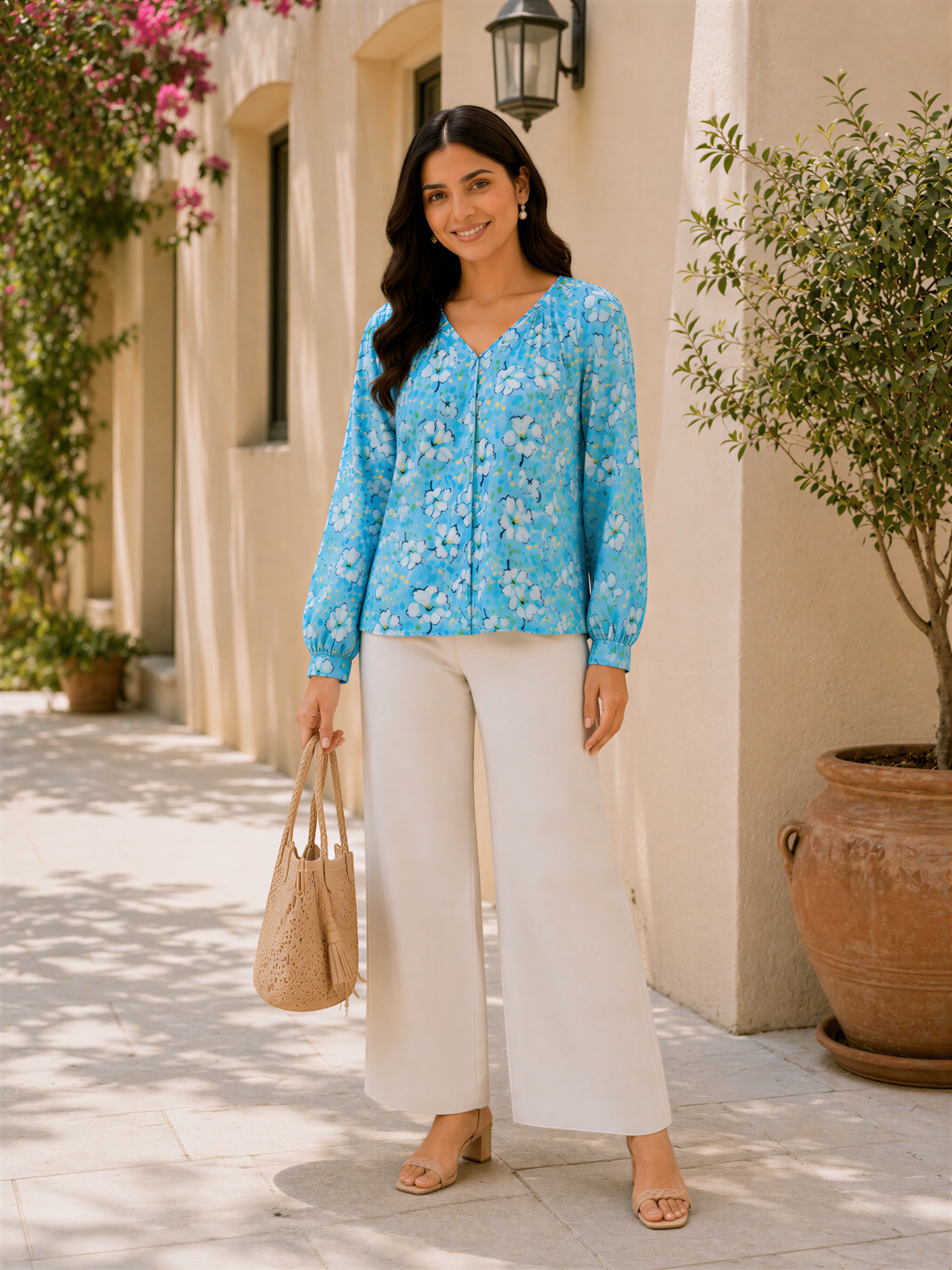 A woman wearing a blue floral blouse and white pants stands on a sidewalk, smiling and holding a beige handbag.