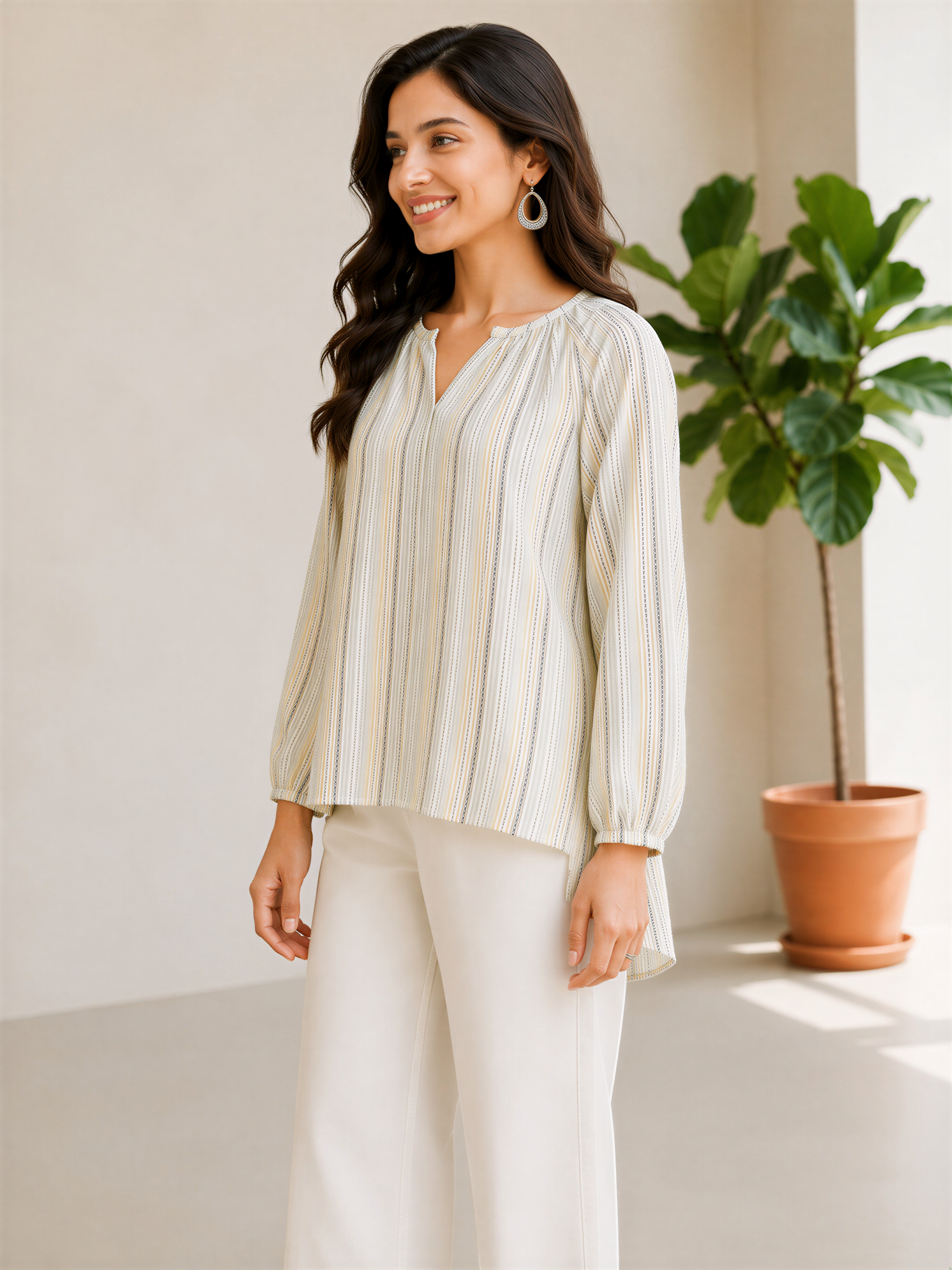 A woman with long dark hair is standing in a room, smiling and wearing a striped blouse and white pants.