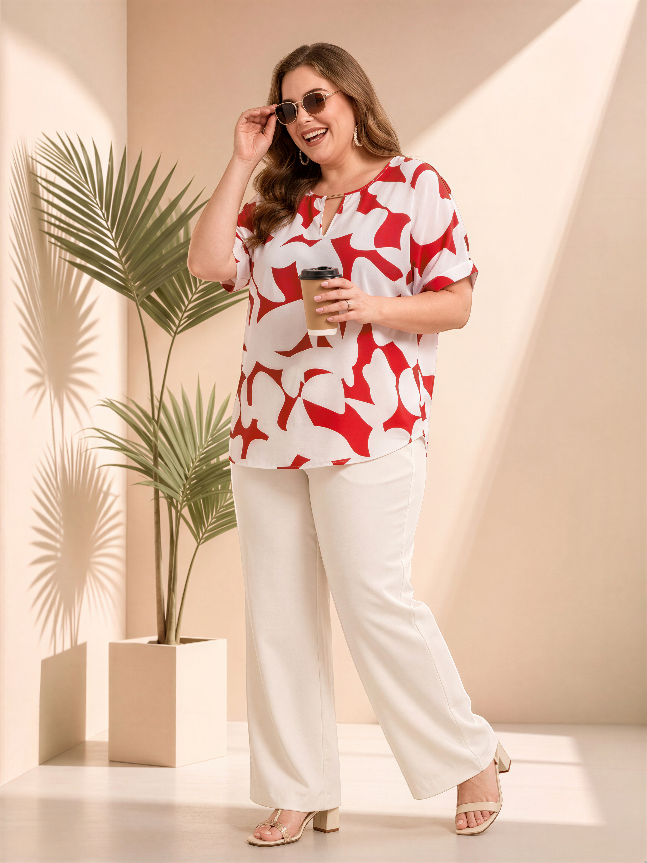A woman wearing a red and white patterned blouse and white pants stands in a room with a potted plant, smiling and holding a coffee cup.