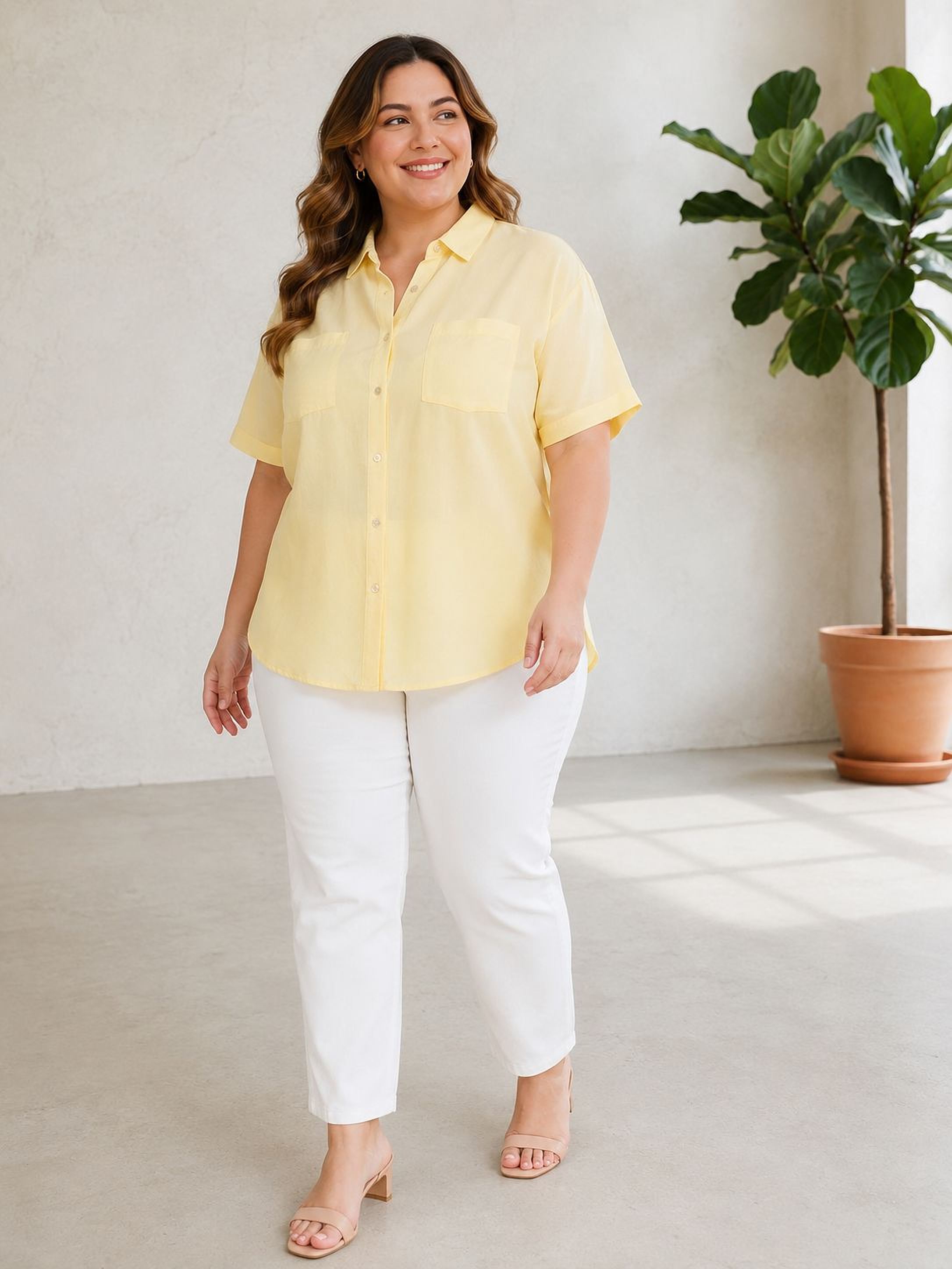 A woman wearing a yellow shirt and white pants is standing in a room with a potted plant in the background.