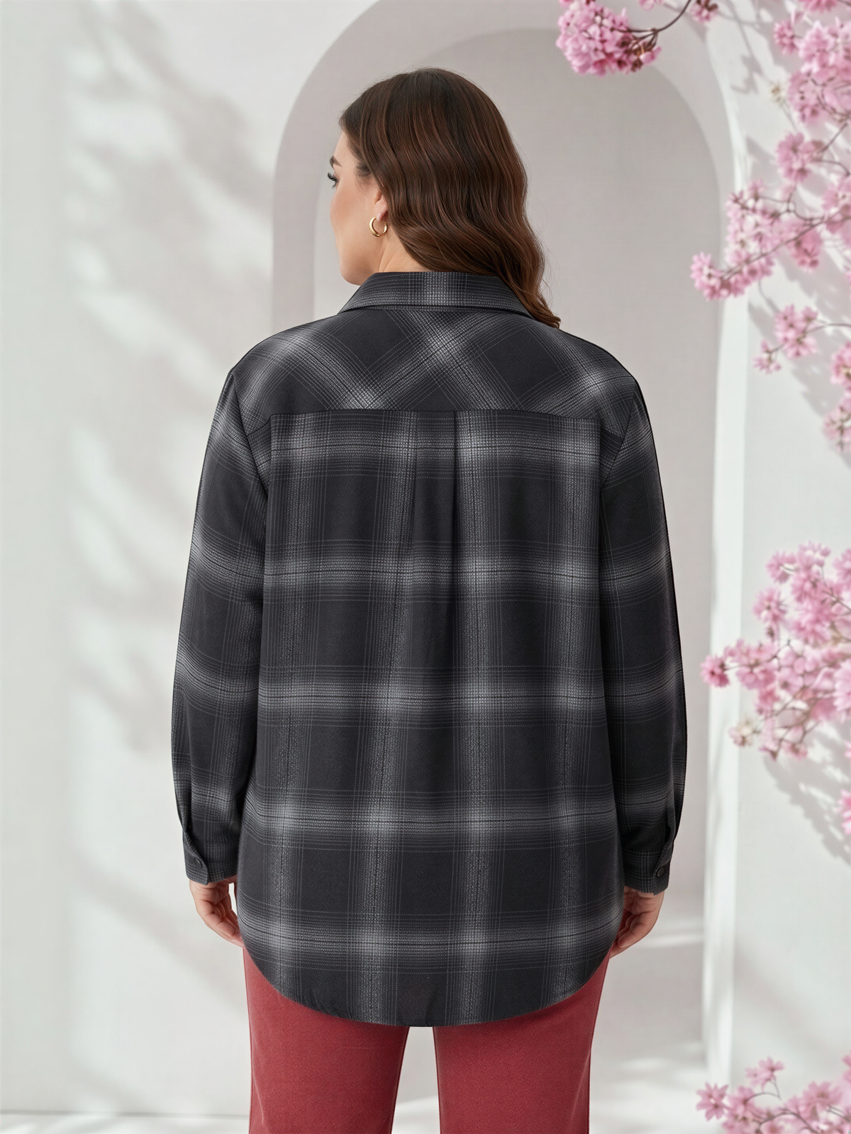 A woman wearing a grey plaid shirt stands in front of a white archway with pink flowers.