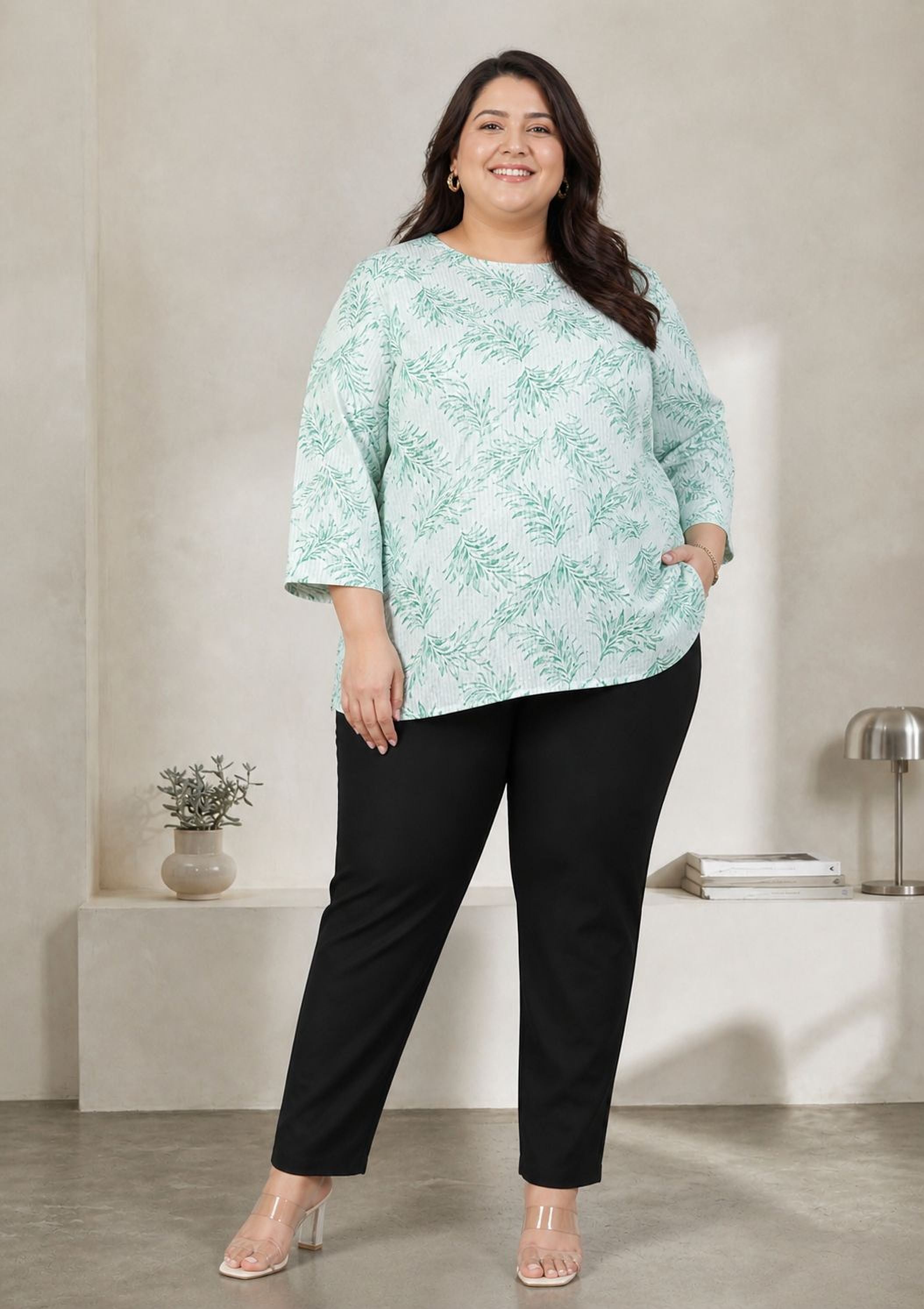 A woman wearing a green and white floral top and black pants stands in a room with a minimalist design.