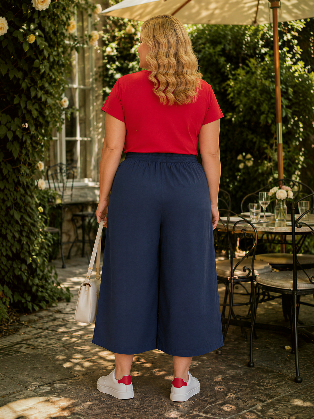 A woman wearing a red top and navy blue wide-leg pants stands in front of a table with a vase of flowers and a white purse.