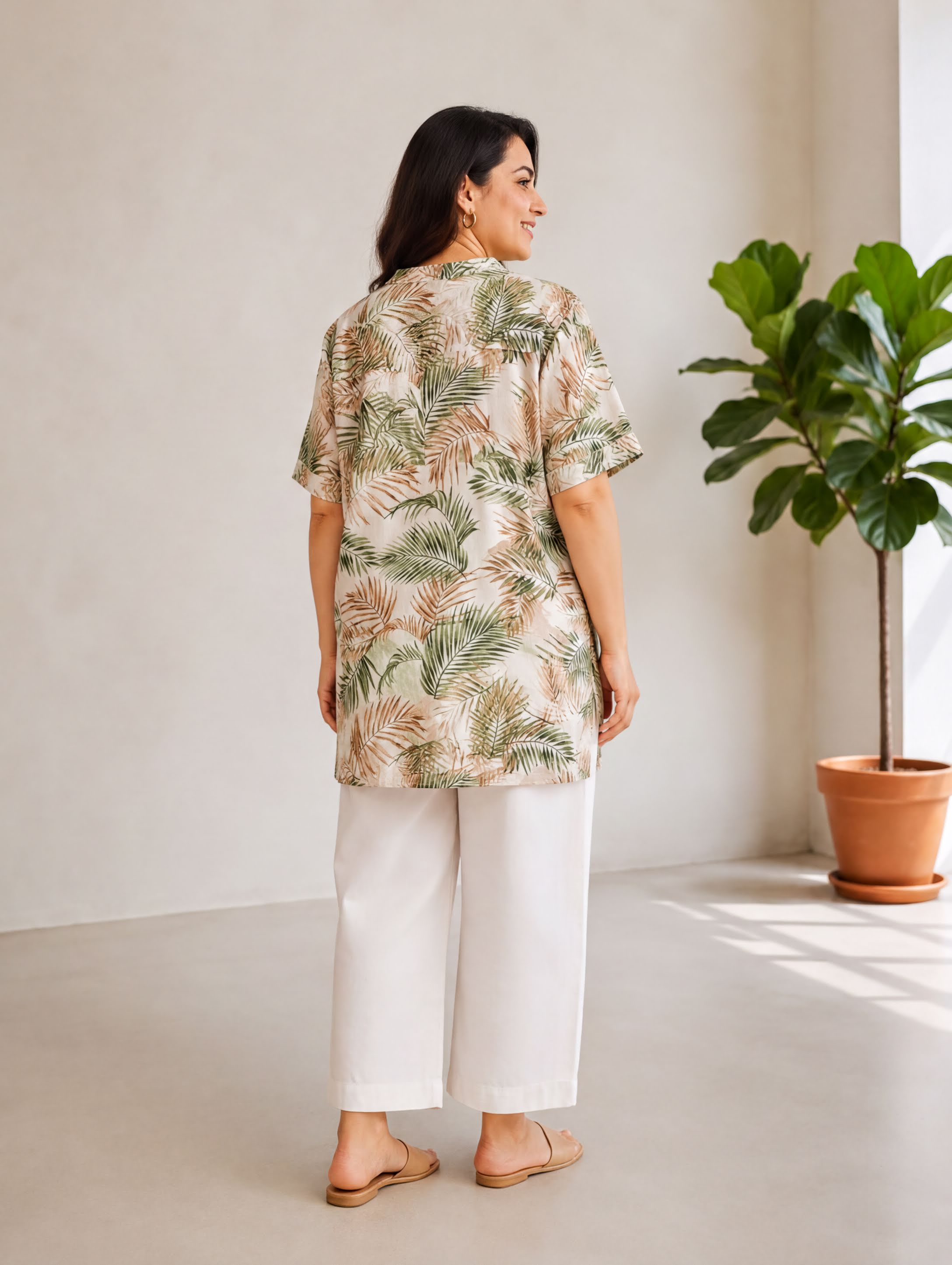 A woman wearing a floral print blouse and white pants stands in a room with a potted plant in the background.
