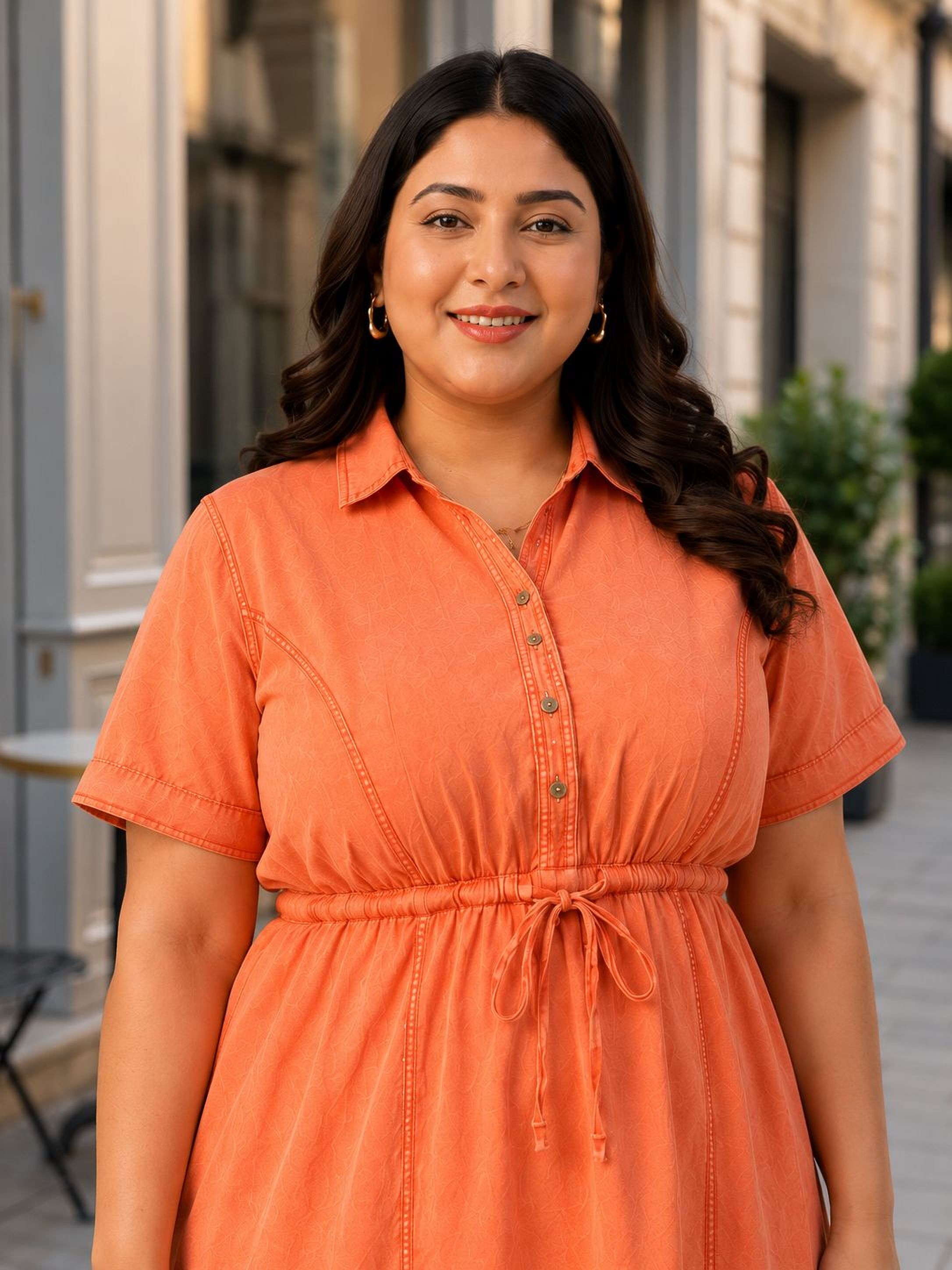 A woman with long dark hair is smiling and wearing an orange dress with a drawstring waist.