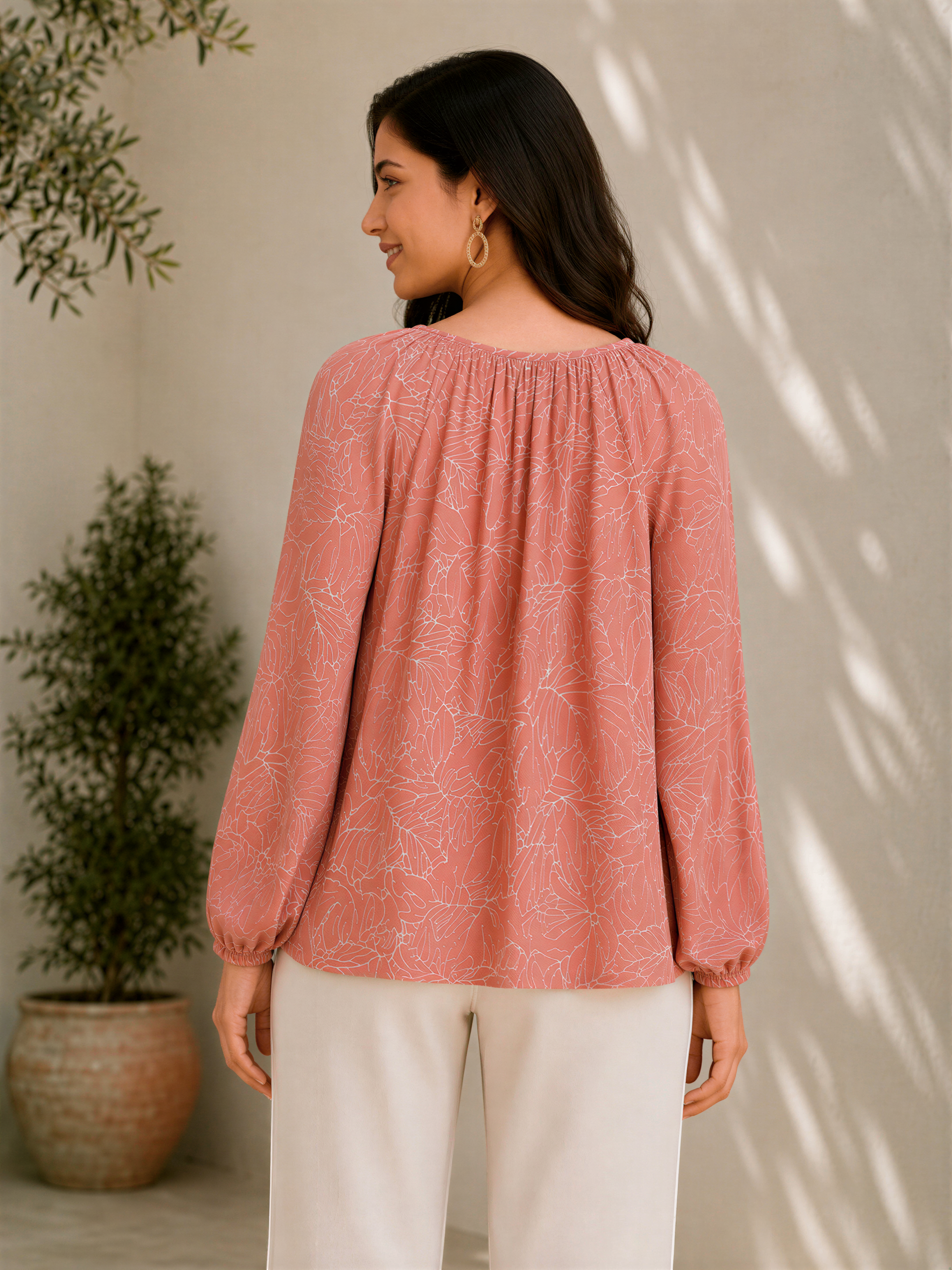 A woman wearing a pink blouse with a leaf pattern stands in front of a white wall, with a potted plant to her left.