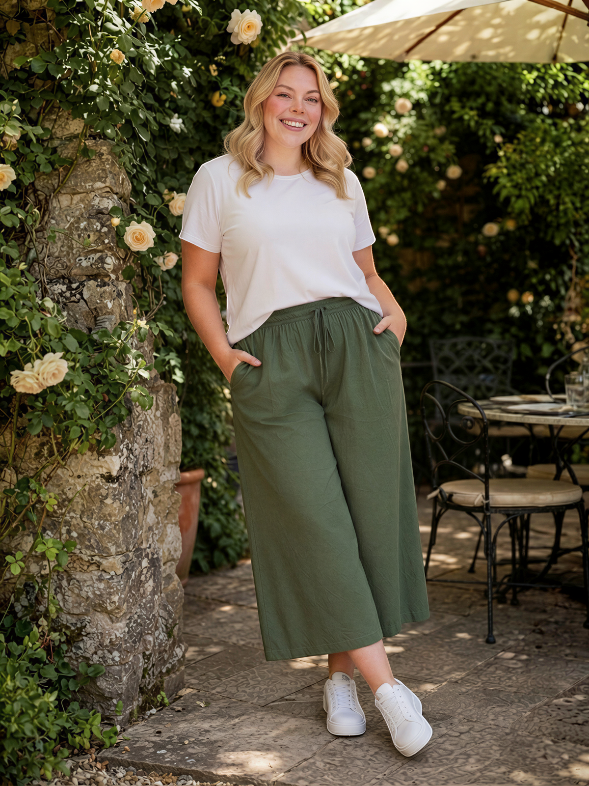 A woman in a white shirt and olive green wide-leg pants stands in front of a stone wall with flowers, smiling and leaning against it.