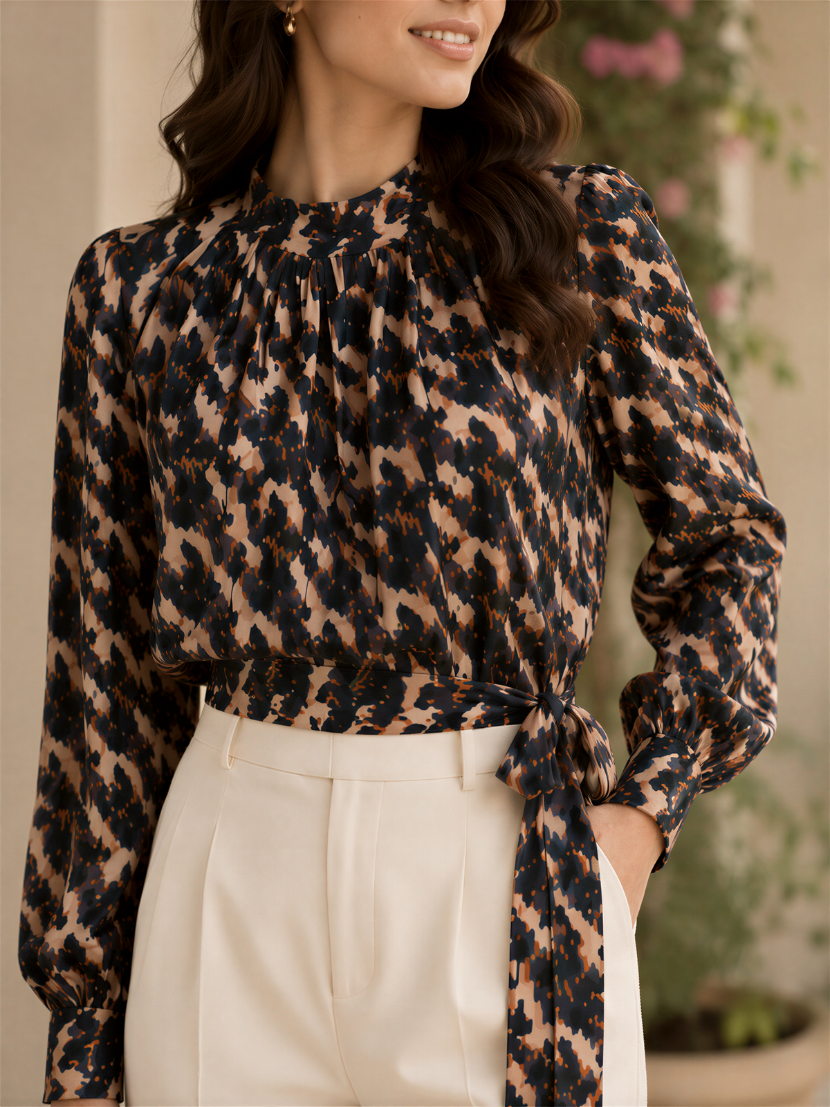 A woman wearing a black and brown patterned blouse with a high neck and long sleeves, paired with white pants.