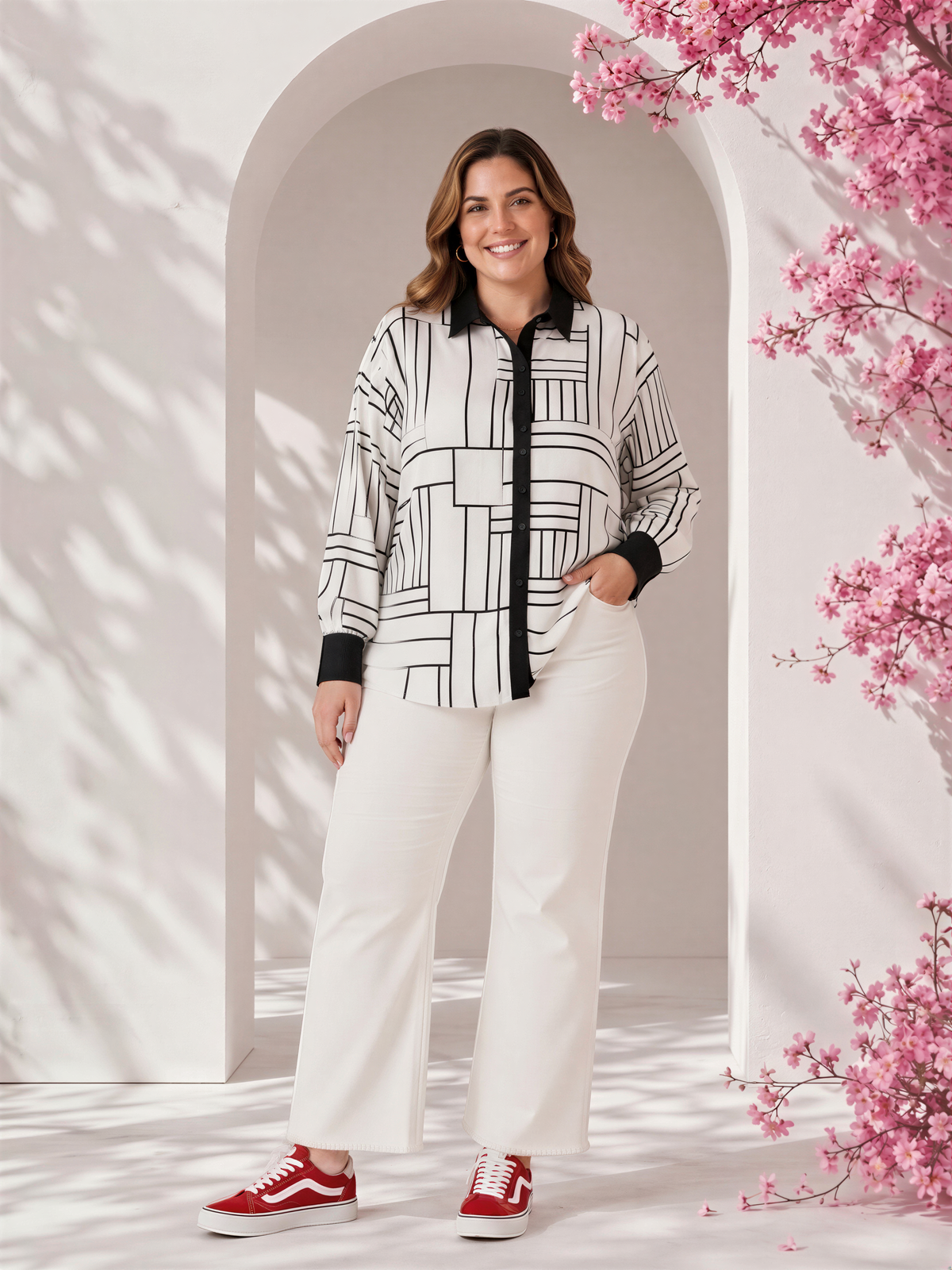 A woman wearing a black and white patterned shirt and white pants stands in front of a white archway with pink flowers.