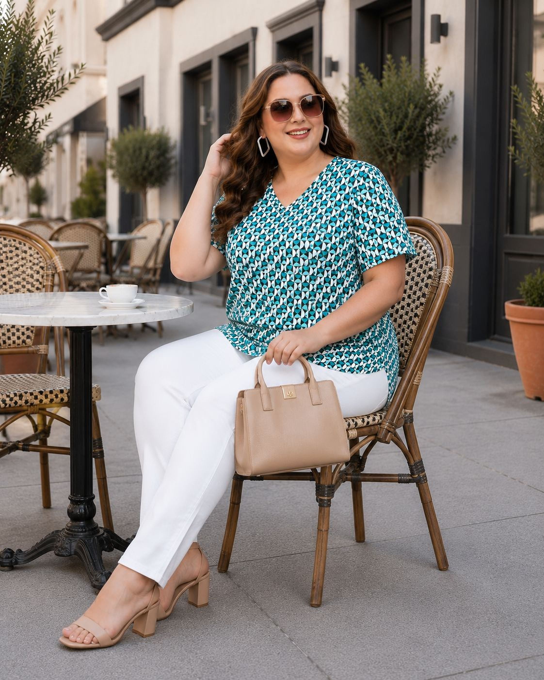 A woman wearing a green and white patterned blouse and white pants is sitting at an outdoor table, smiling and holding a tan handbag.