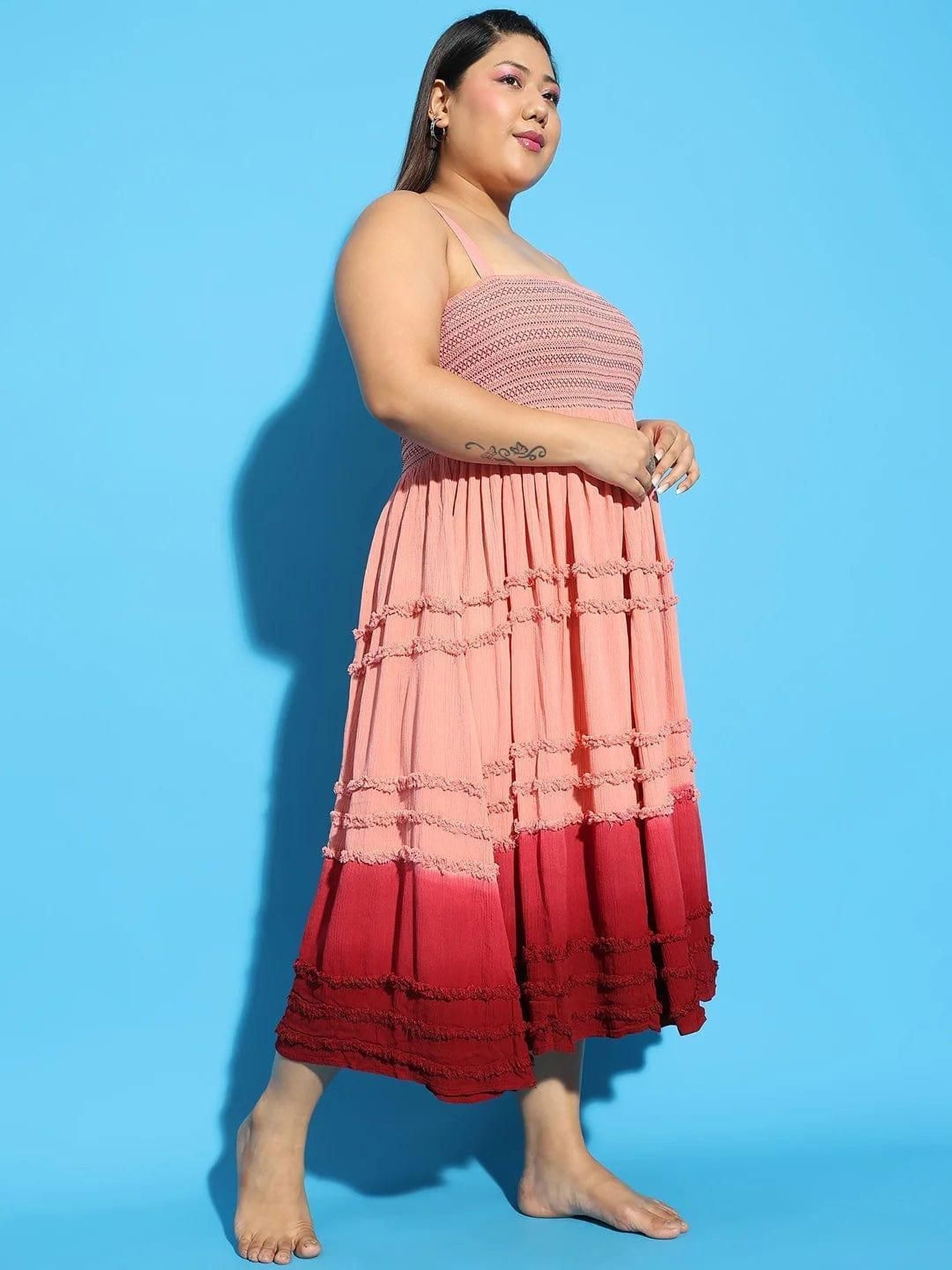 A woman wearing a pink and red tiered dress and a wide-brimmed hat is smiling and gesturing with her hand.