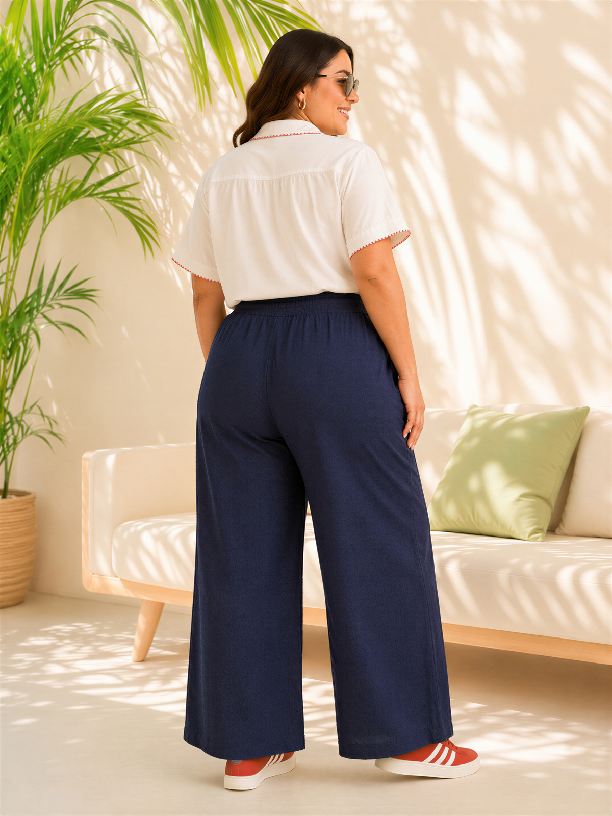 A woman wearing a white blouse and navy blue wide-leg pants stands in a room with a white couch and a potted plant.