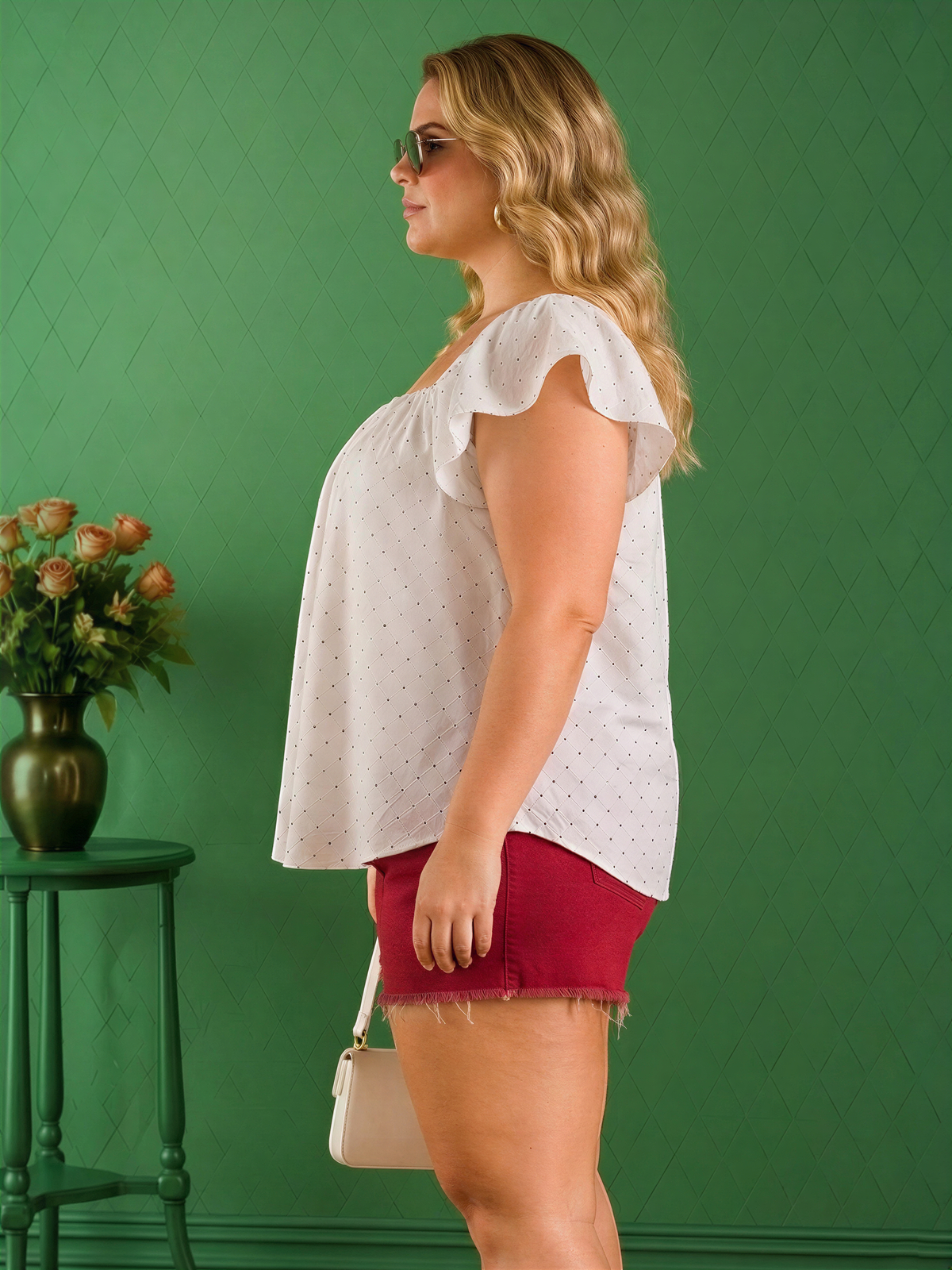A woman with blonde hair wearing a white polka dot blouse and red shorts stands in front of a green wall, holding a white purse.