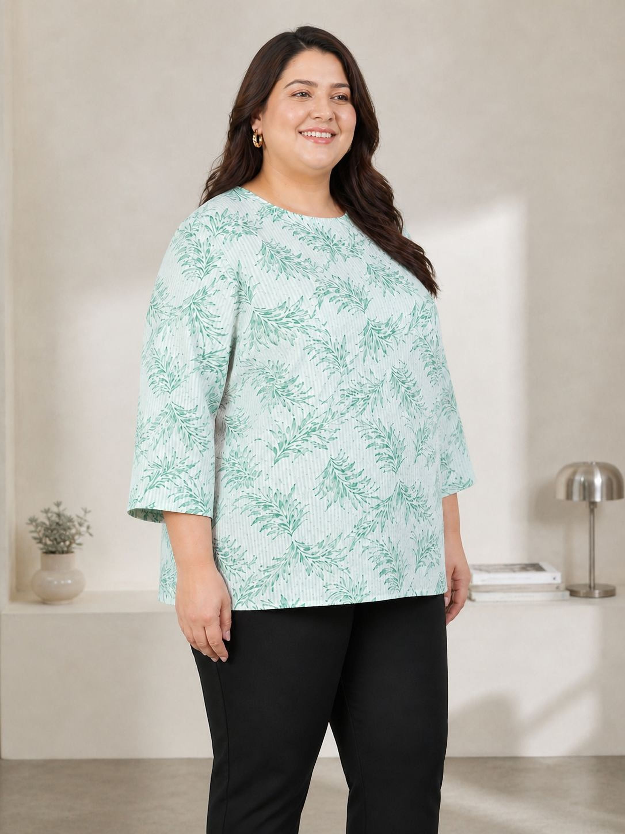 A woman wearing a green and white patterned blouse stands in a room with a lamp and a plant in the background.