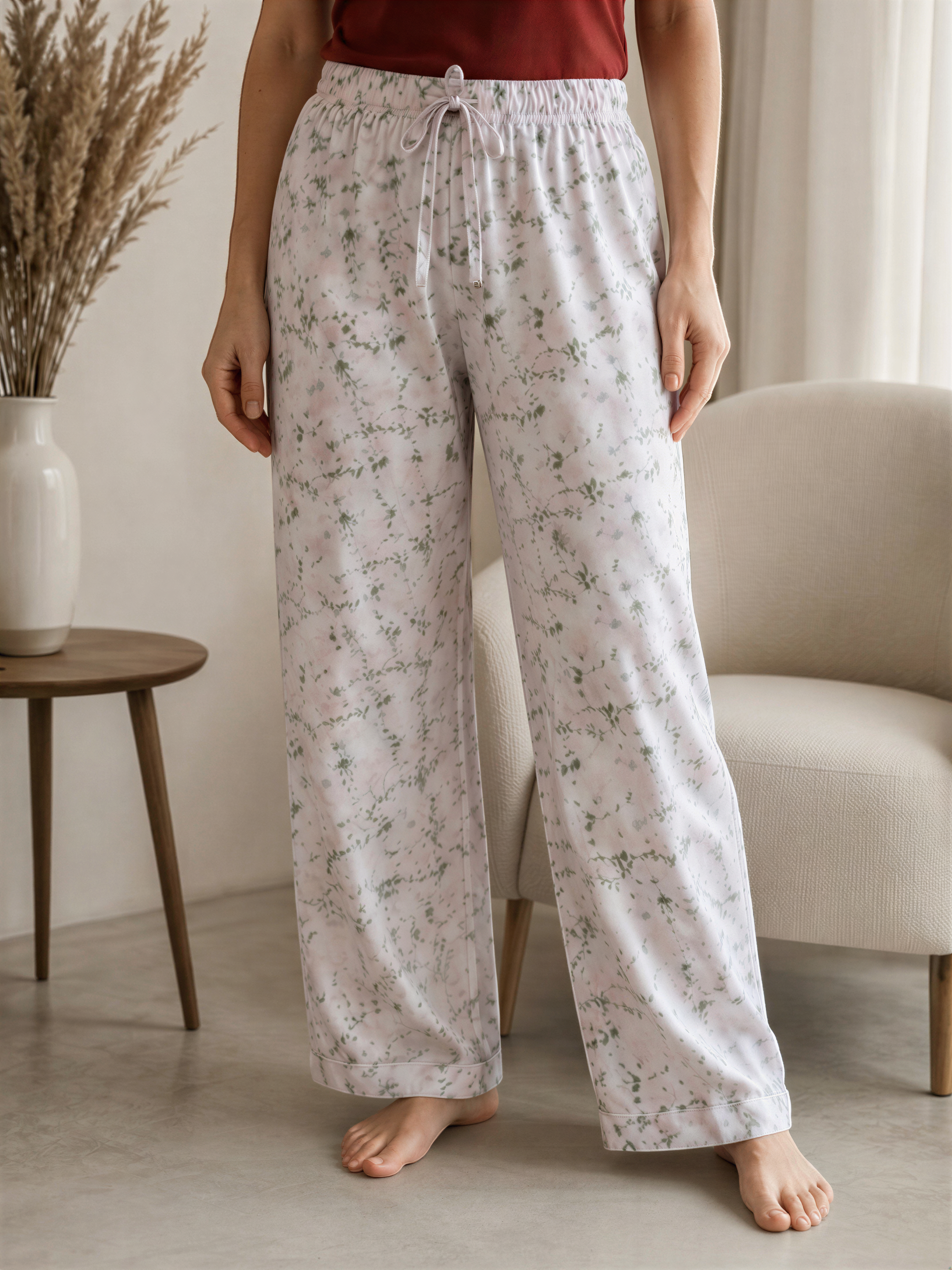 A person wearing white floral pajama pants stands in a room with a vase of dried flowers and a beige armchair.