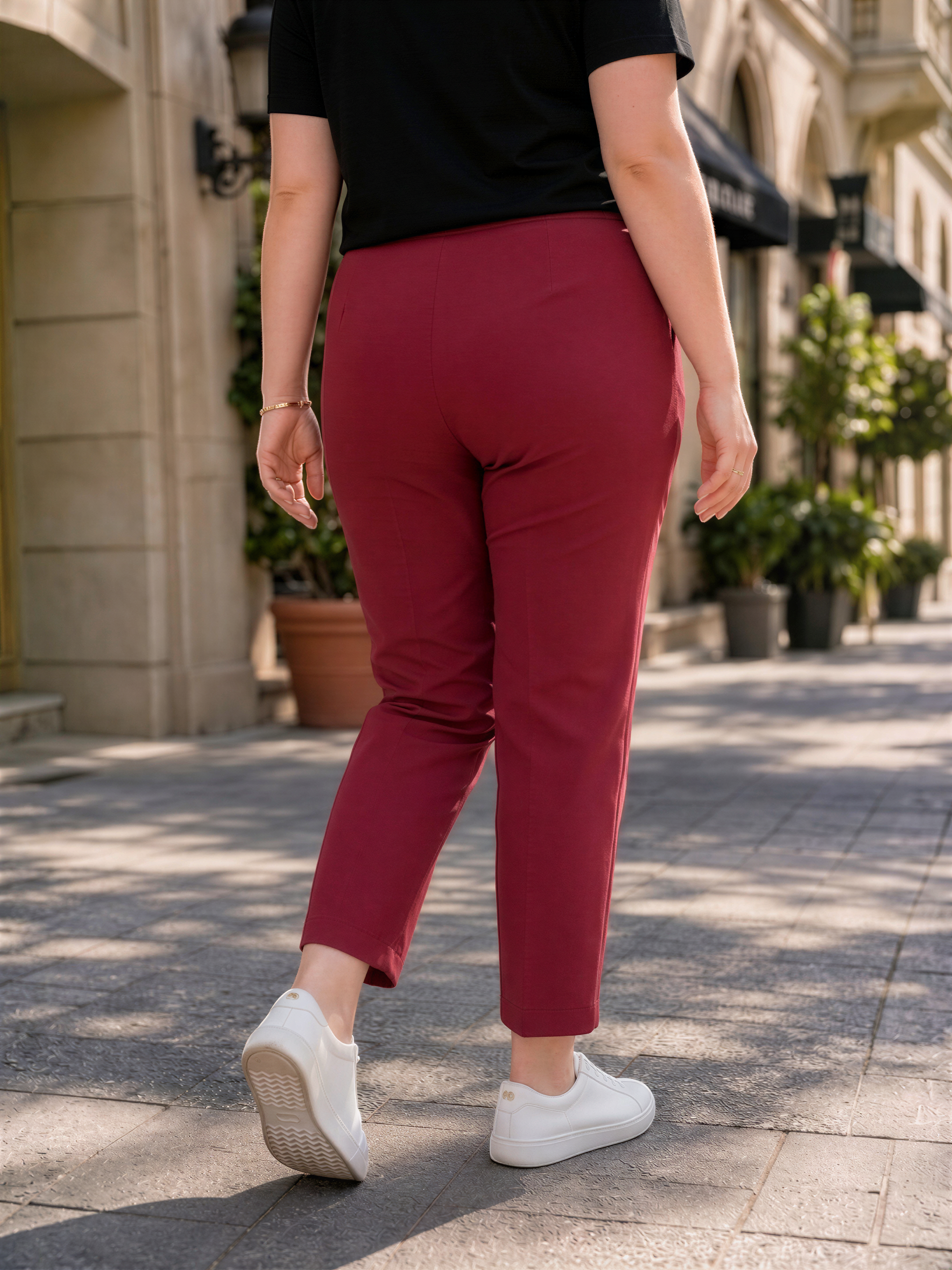 A person wearing a black shirt and red pants is walking on a sidewalk, with potted plants and a building in the background.