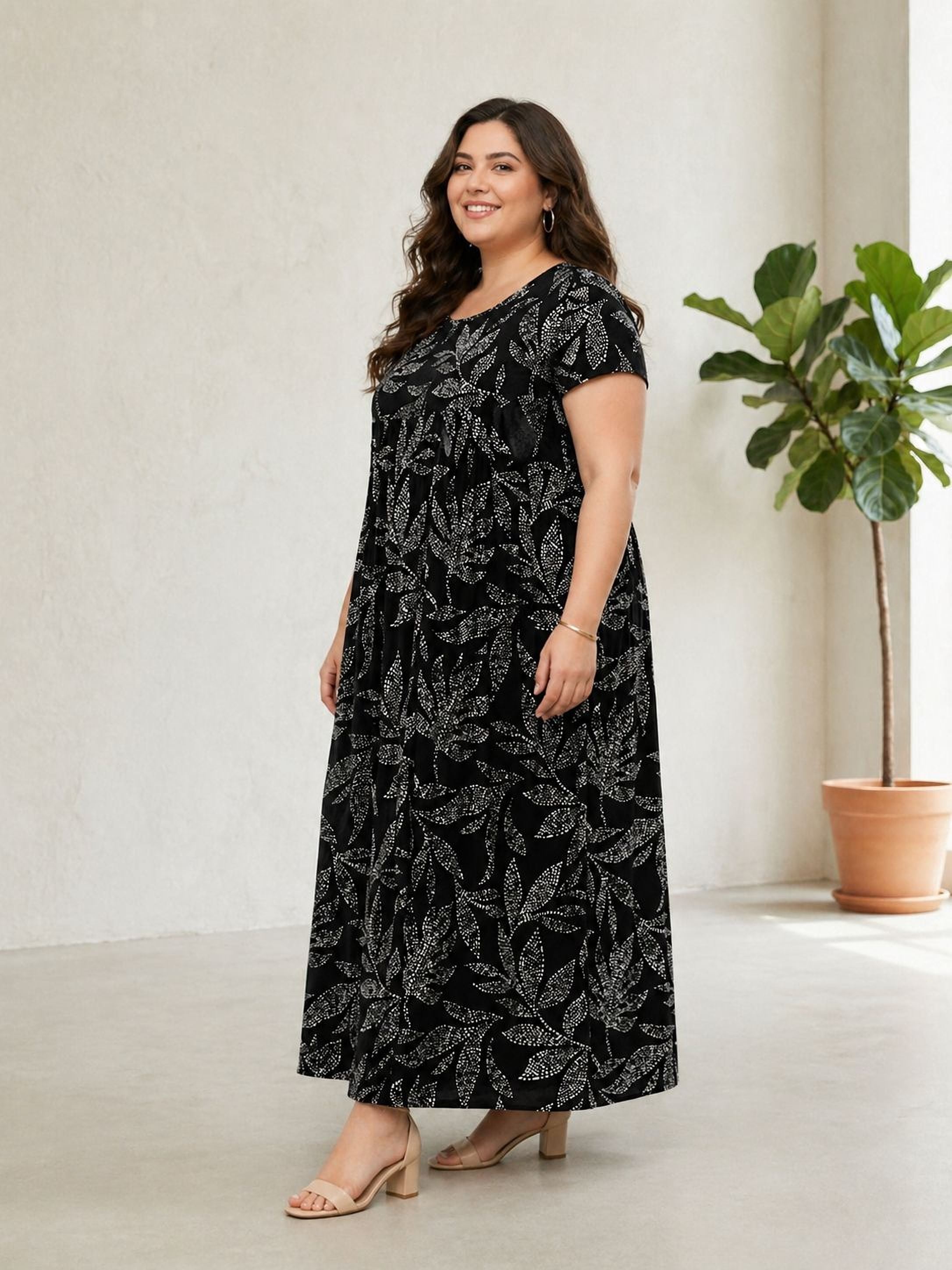 A woman wearing a long black dress with a floral pattern stands in a room with a potted plant in the background.