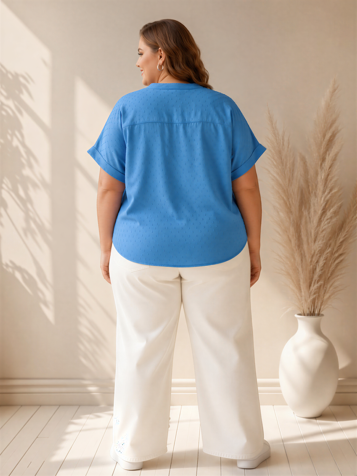 A woman wearing a blue blouse and white pants stands in a room with a potted plant to her right.