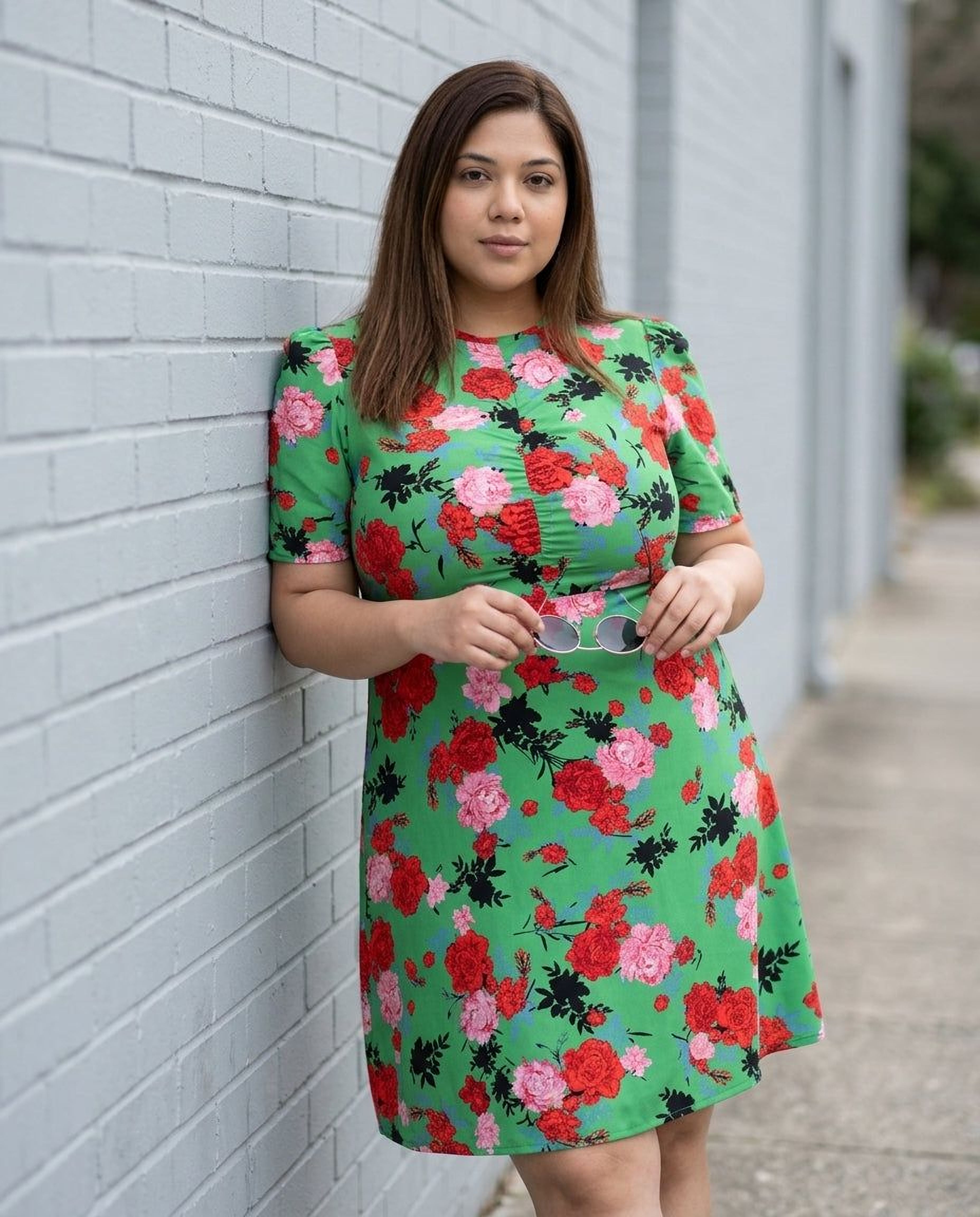 A woman wearing a green floral print dress and sunglasses, holding a black purse, and smiling at the camera.
