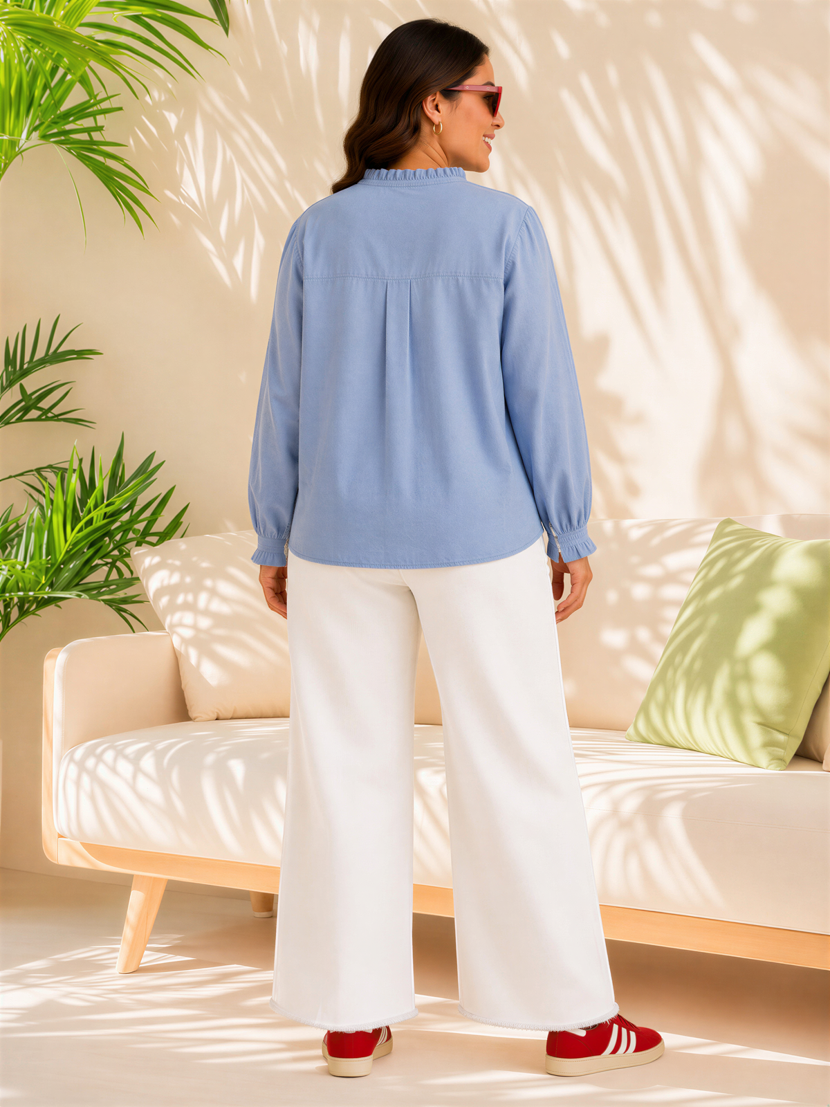 A woman wearing a blue blouse and white pants stands in front of a couch with a green pillow, with a potted plant in the background.