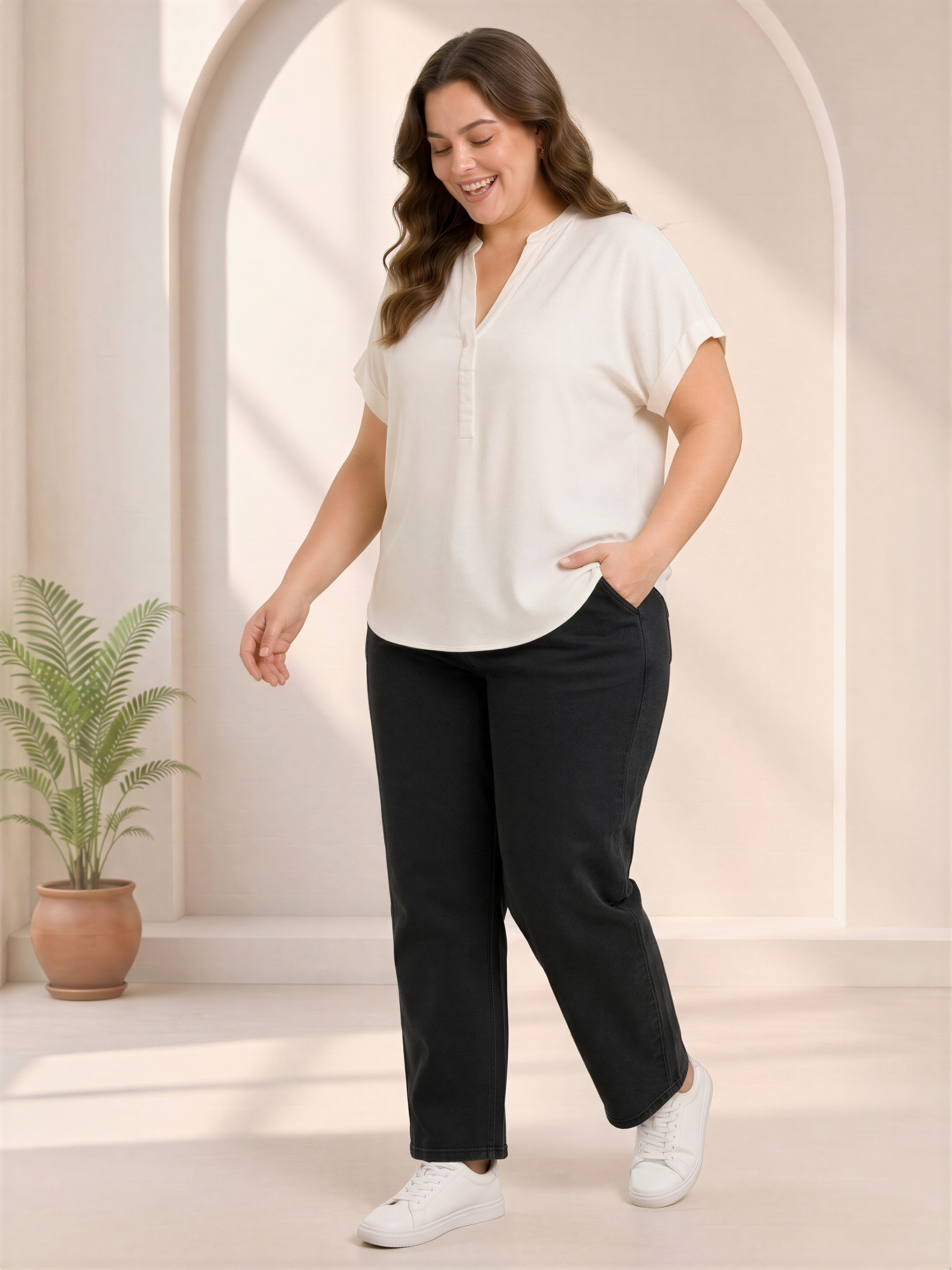 A woman in a white blouse and black pants stands in a room with a potted plant in the background.