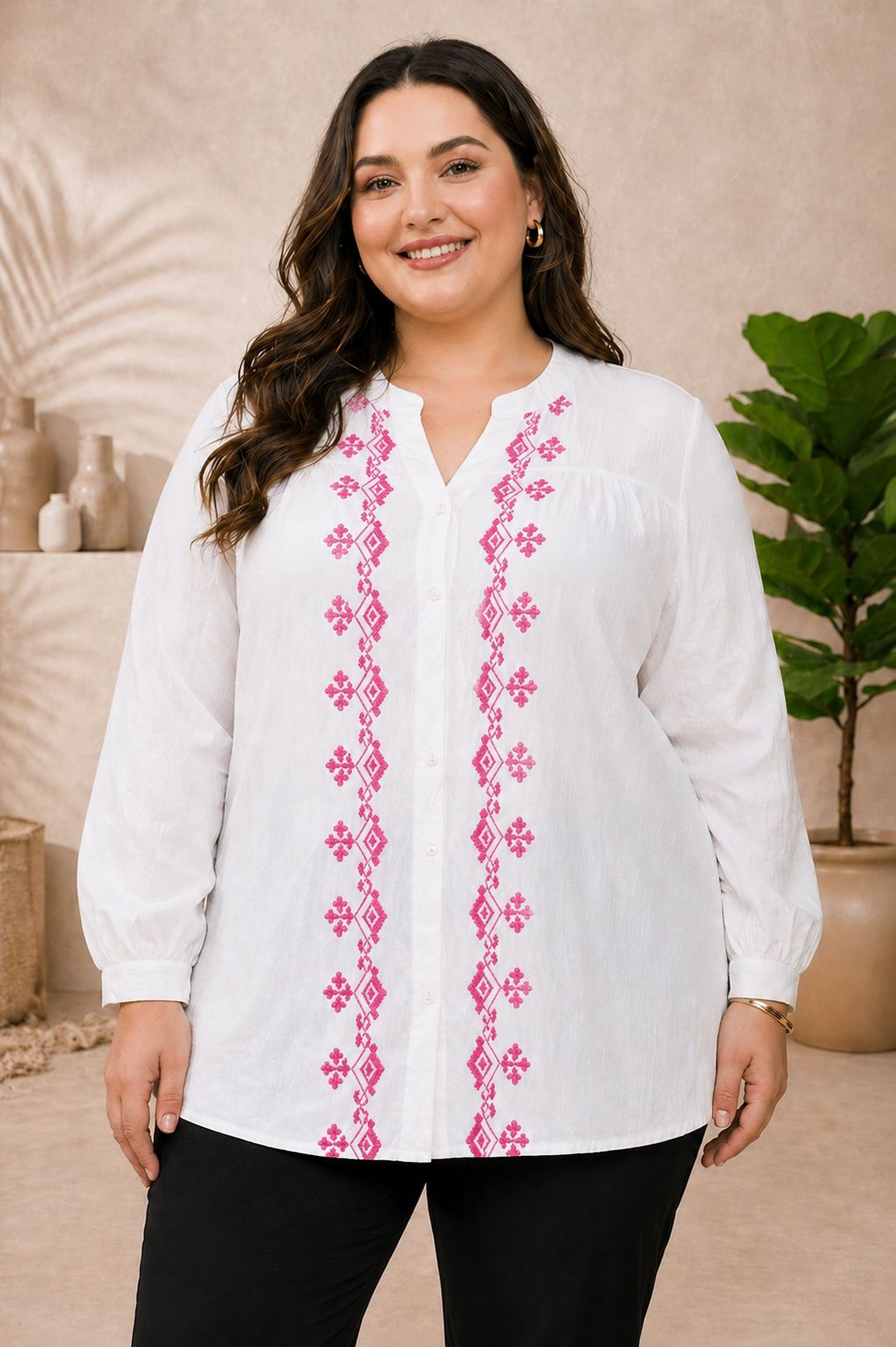 A woman wearing a white blouse with pink embroidered patterns stands in front of a beige wall, smiling at the camera.
